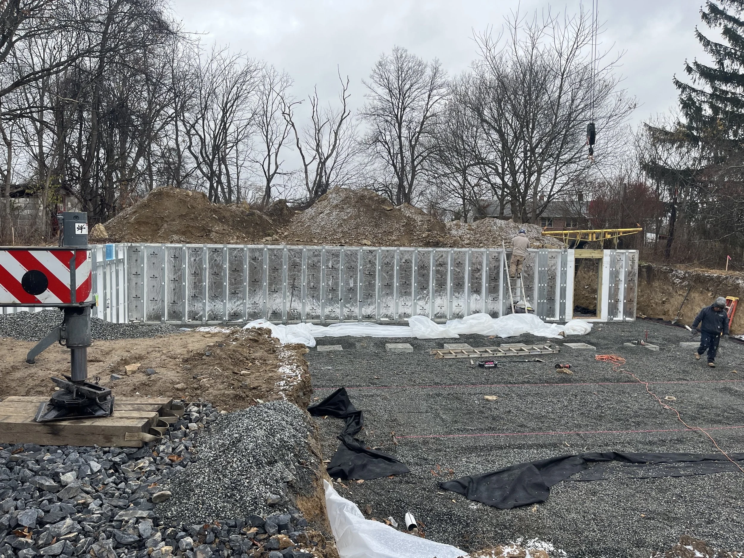 Construction site featuring workers installing or repairing a concrete retaining wall with gravel and soil piles in the background, leafless trees, and construction equipment.