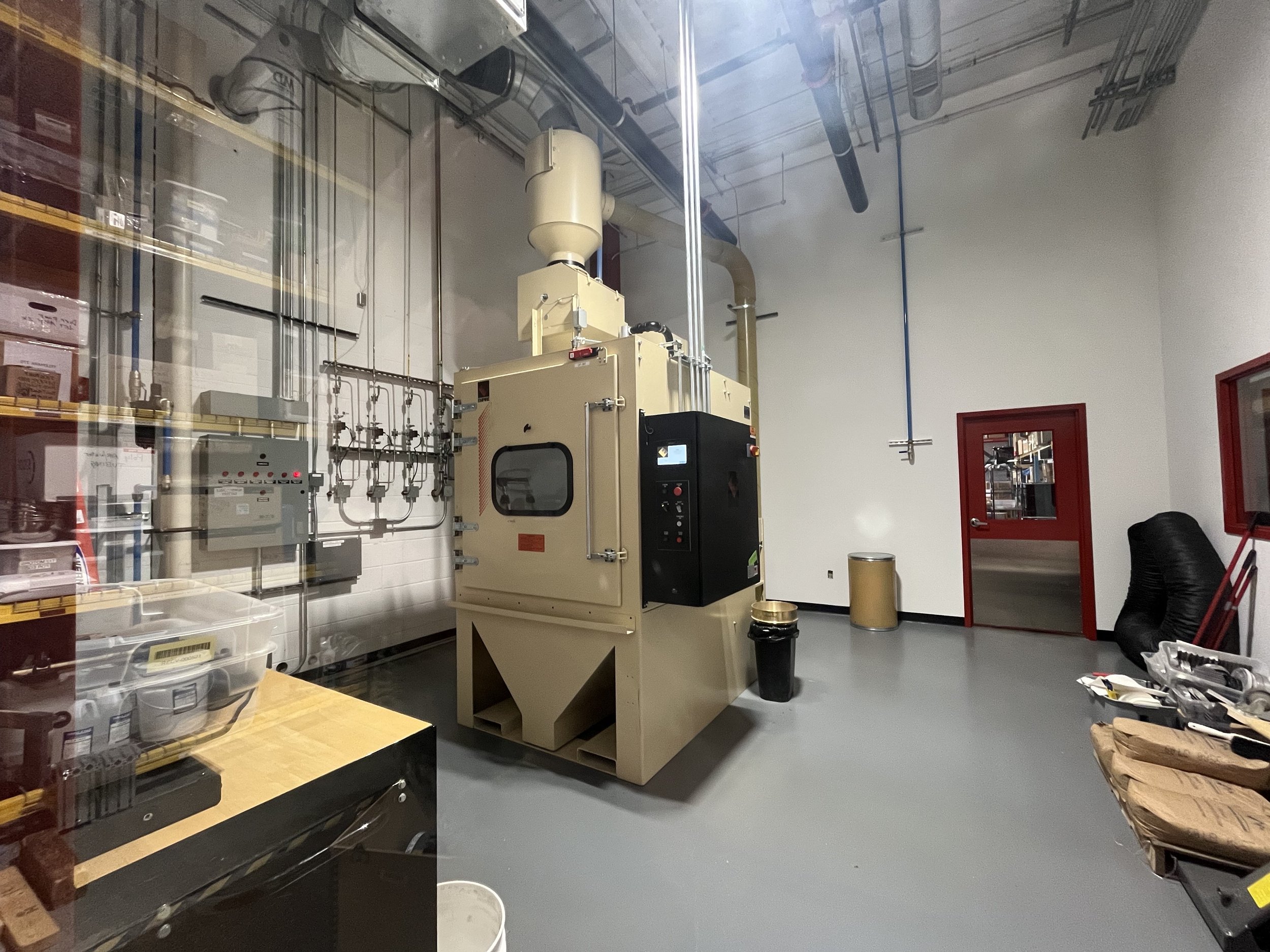 Industrial laboratory with a large beige machine, various pipes and control panels, a workbench with tools, and a red door leading to another room.