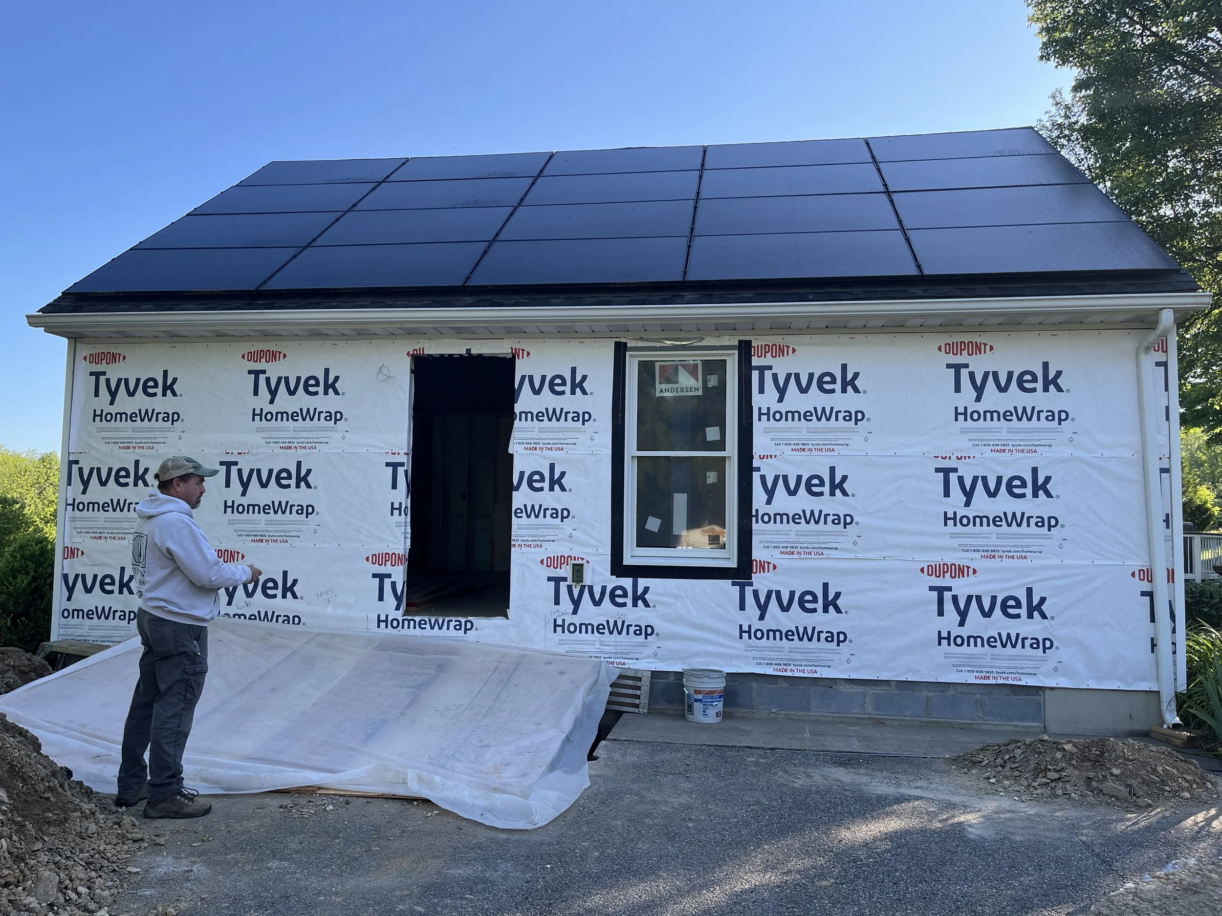 A house under construction with Tyvek HomeWrap, black roofing panels, and a window. A man is standing nearby holding a mobile phone.