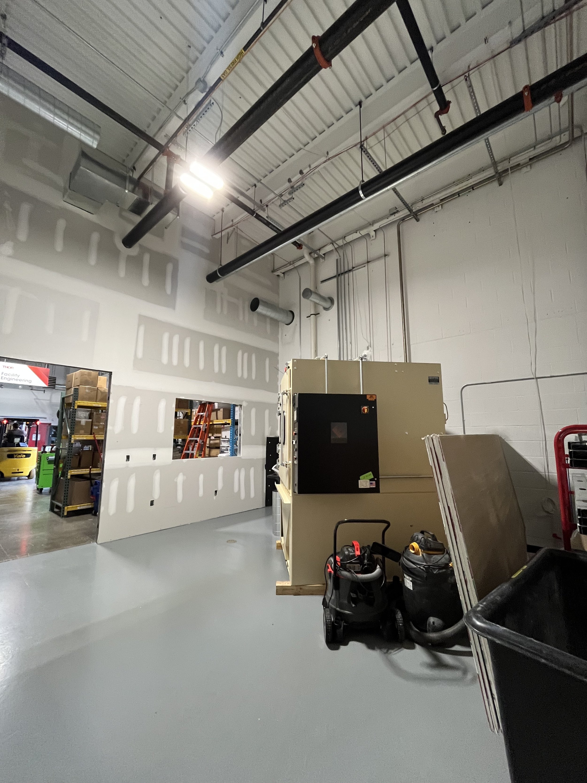 View of an industrial or commercial workspace with construction or maintenance equipment, including a large beige machine, two wet/dry vacuums, and various tools, against white unfinished walls with visible drywall. Overhead, black pipes and ducts ru