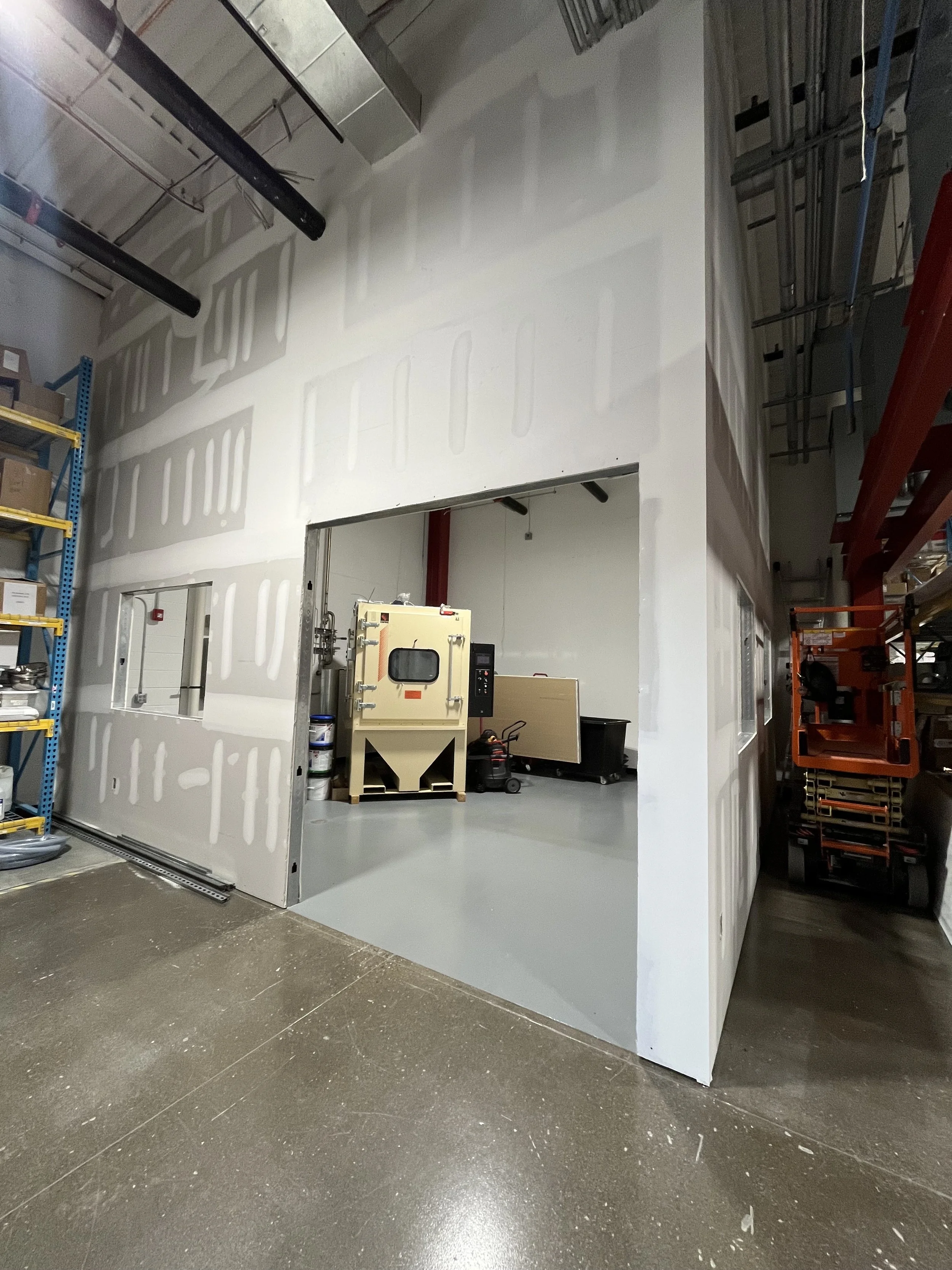 Interior of a warehouse or workshop with an unfinished wall and a large doorway opening to a room with equipment, shelves, and containers. Shelving units and construction tools are visible.