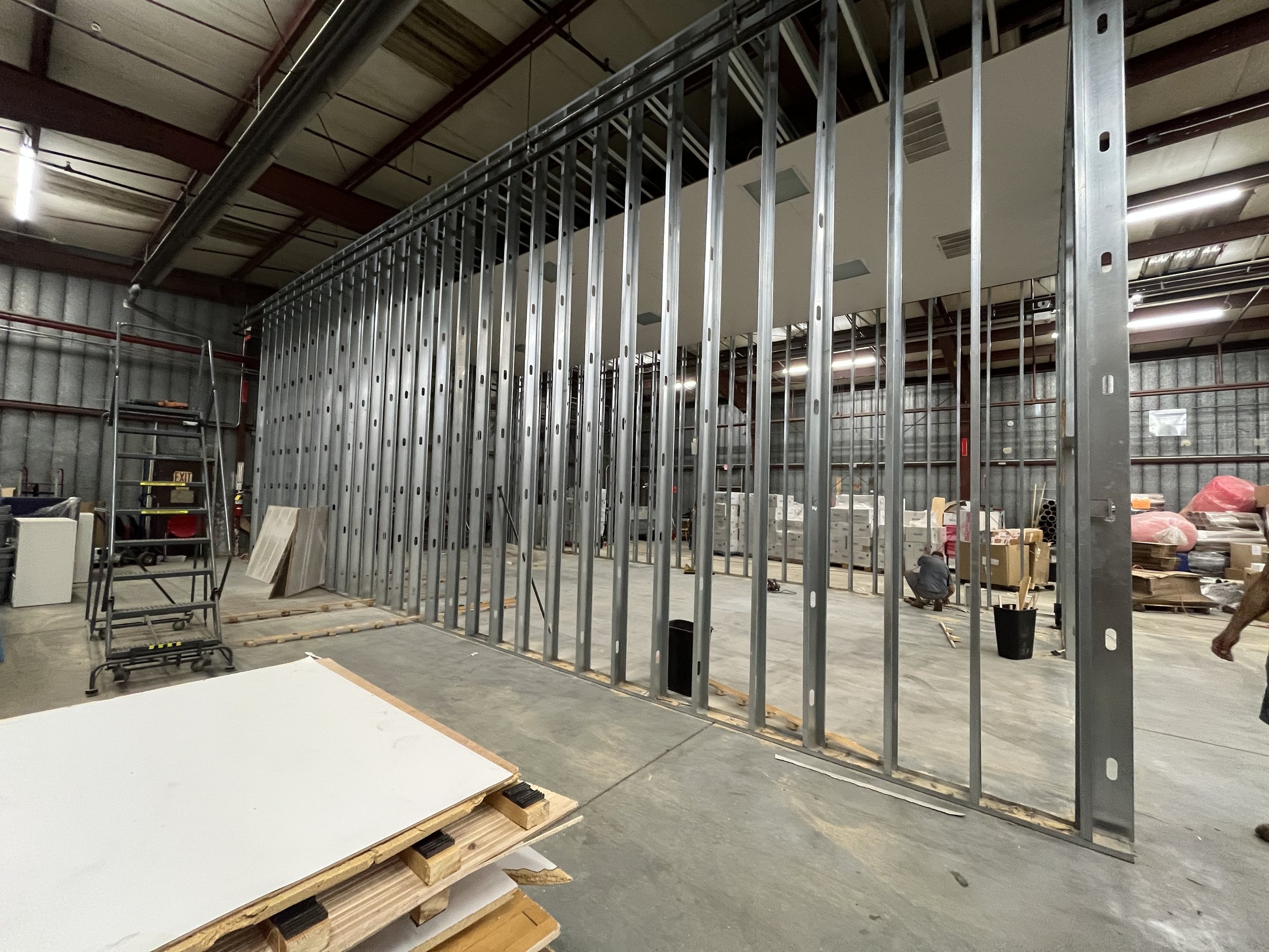 Construction of an interior metal-framed wall in a warehouse or industrial space, with workers installing framing and building materials visible in the background.