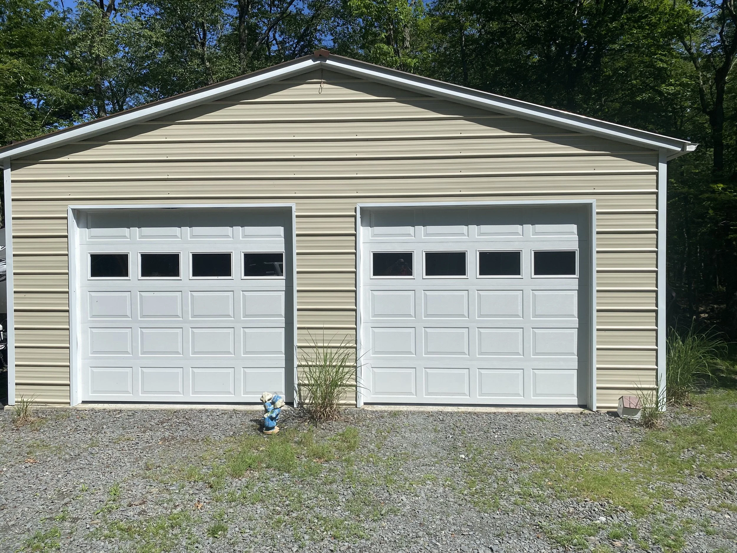 A beige metal garage with two white garage doors, situated on a gravel driveway with some grass and small plants.