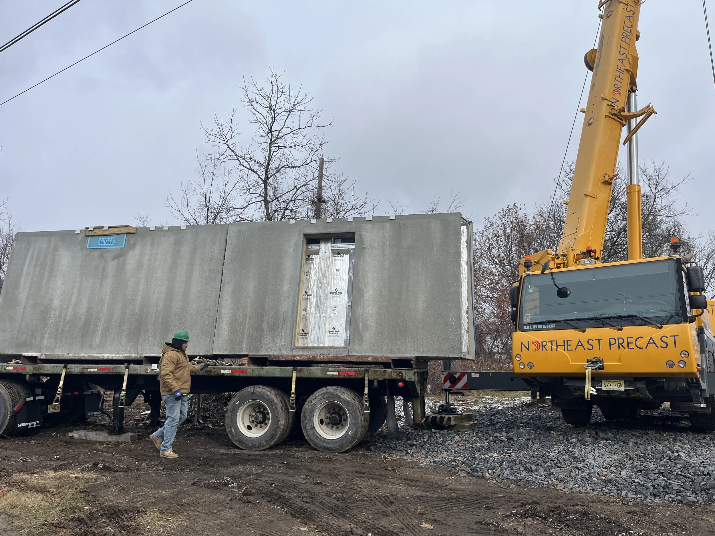 Construction worker standing near a flatbed truck with a large concrete panel and a yellow crane labeled 'NORTHEAST PRECAST' lifting the panel.