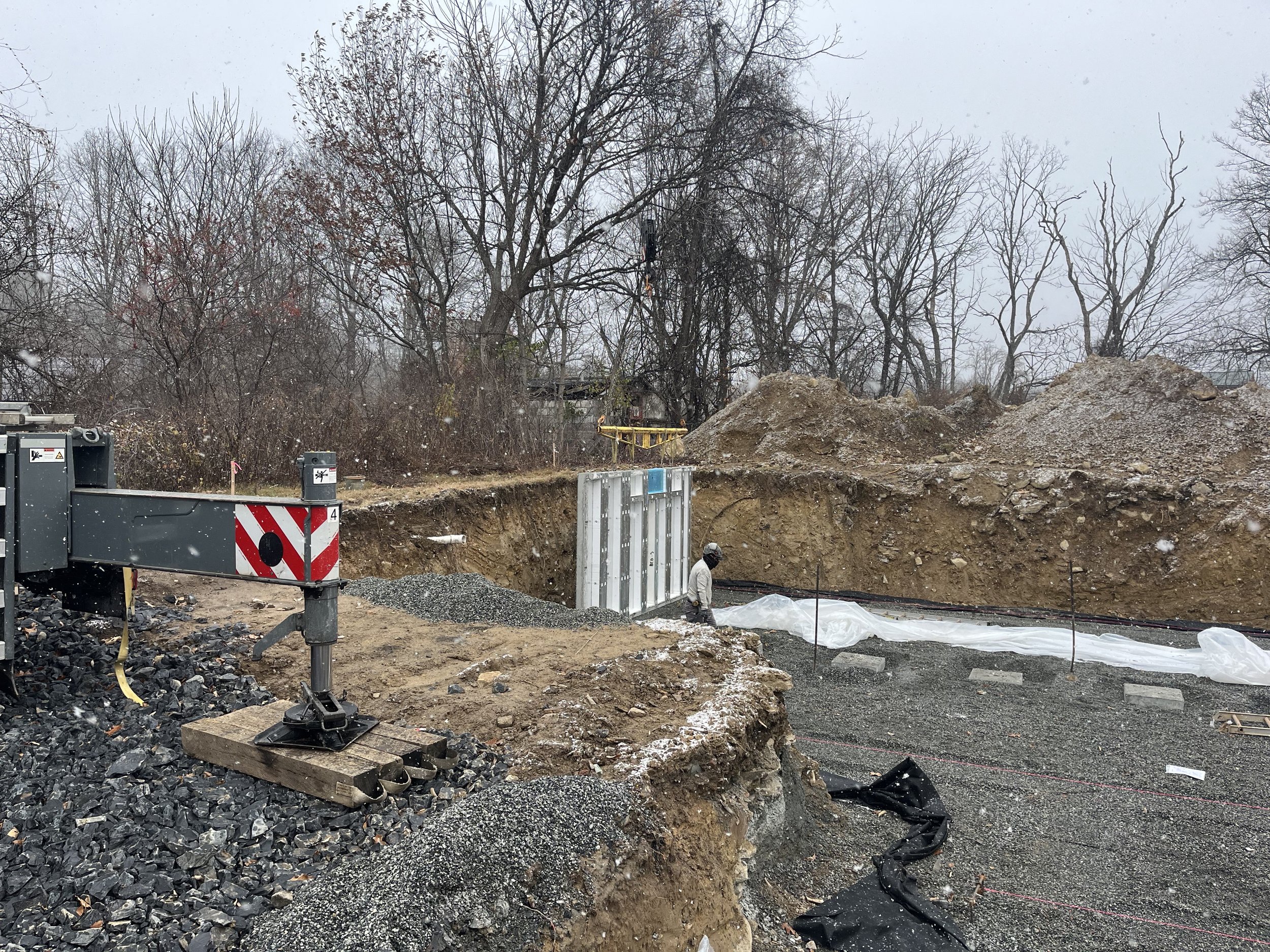 Construction site with a worker in a white jacket and black helmet, excavation area with gravel and soil, and large piles of dirt in the background, overcast sky, leafless trees, and construction equipment.