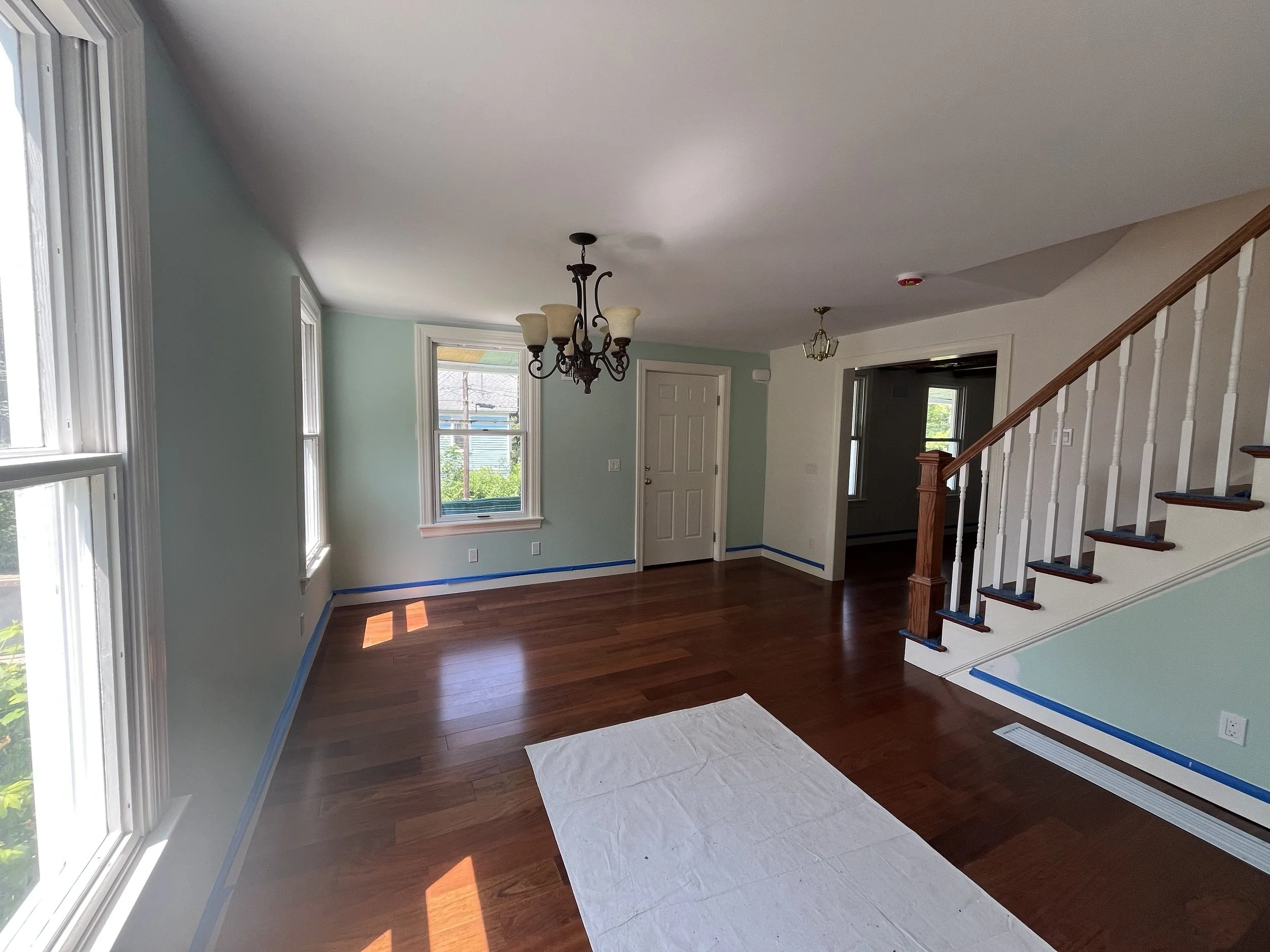 Empty living room with hardwood floors, light green walls, large windows, a staircase with wooden handrail, and a white ceiling. Two chandeliers hang from the ceiling.