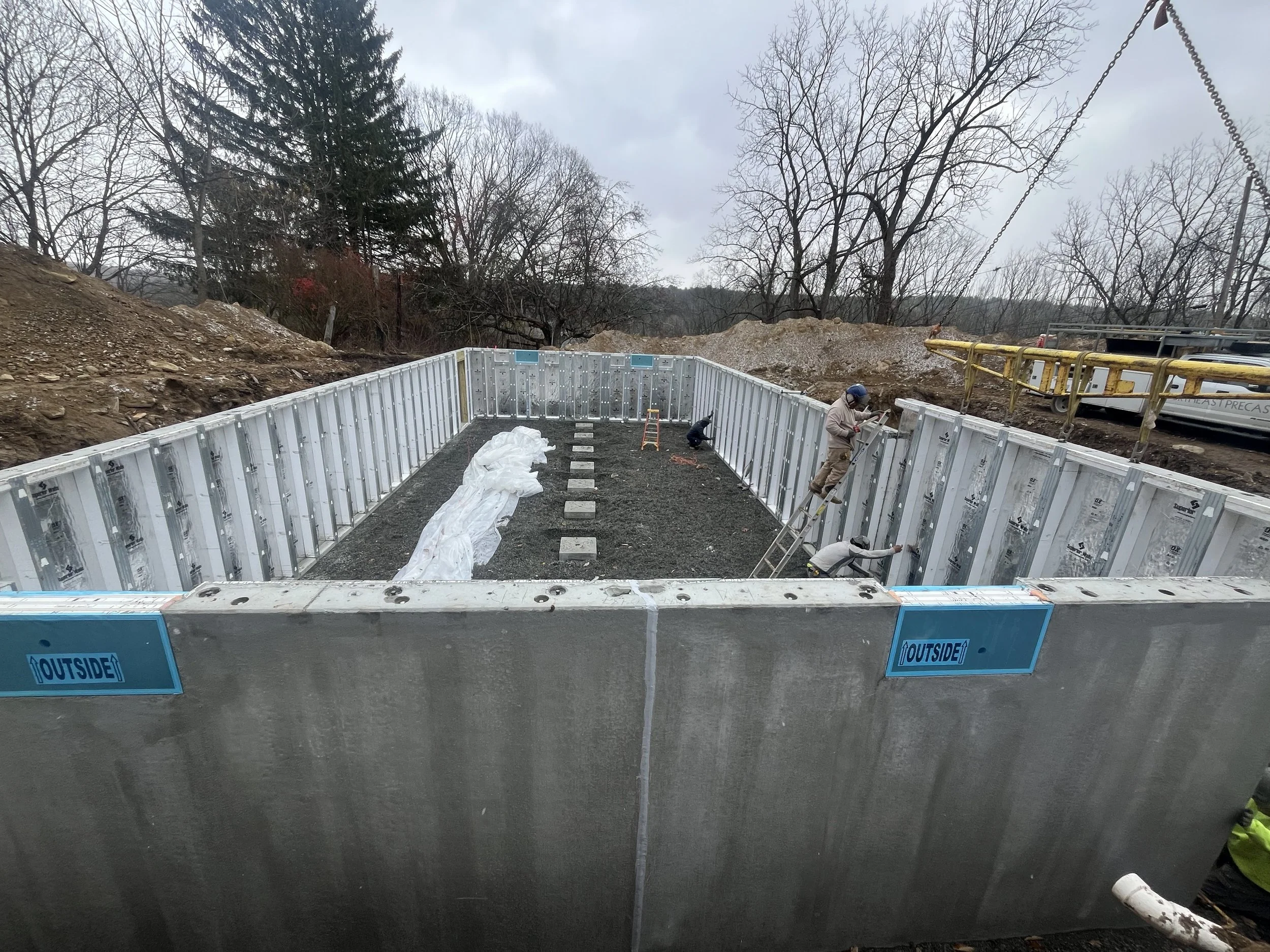 Construction workers assembling a concrete foundation, with metal wall panels and construction equipment on a cloudy day in a wooded area.