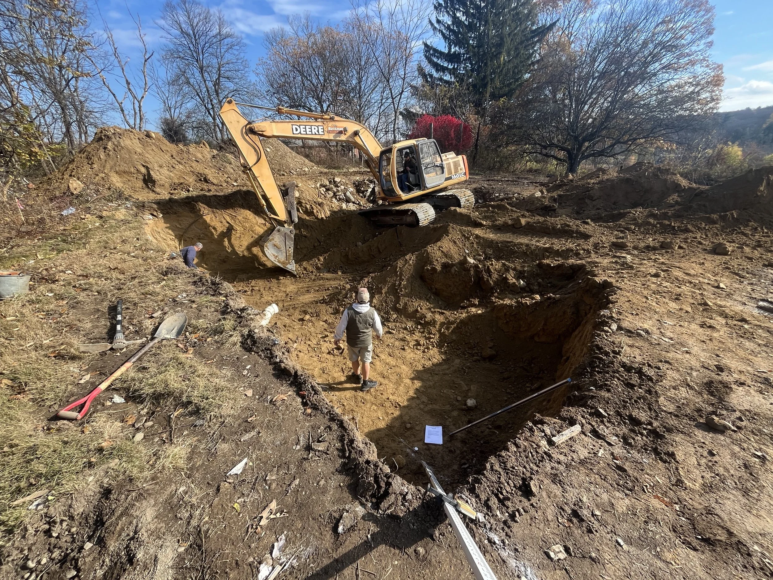 Construction site with a large excavator digging a trench in the ground, and workers nearby assisting with the excavation. There are trees and a clear sky in the background.