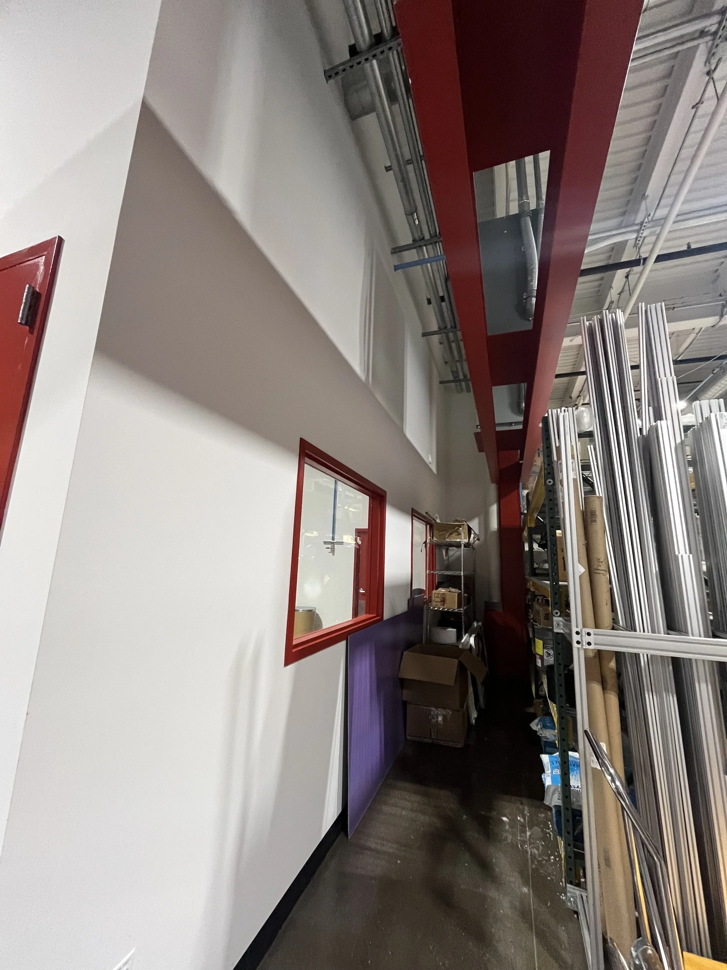 Interior of a warehouse with white and red walls, metal shelving with aluminum poles, boxes, and various materials, with a red-painted beam running across the ceiling and a small office window with a red frame.