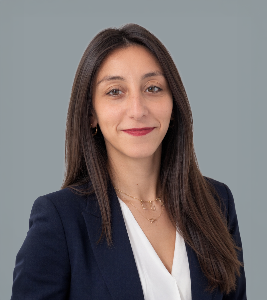 Una donna con capelli castani e occhi castani sorride, indossa una giacca blu, una camicia bianca e gioielli dorati, sfondo grigio.