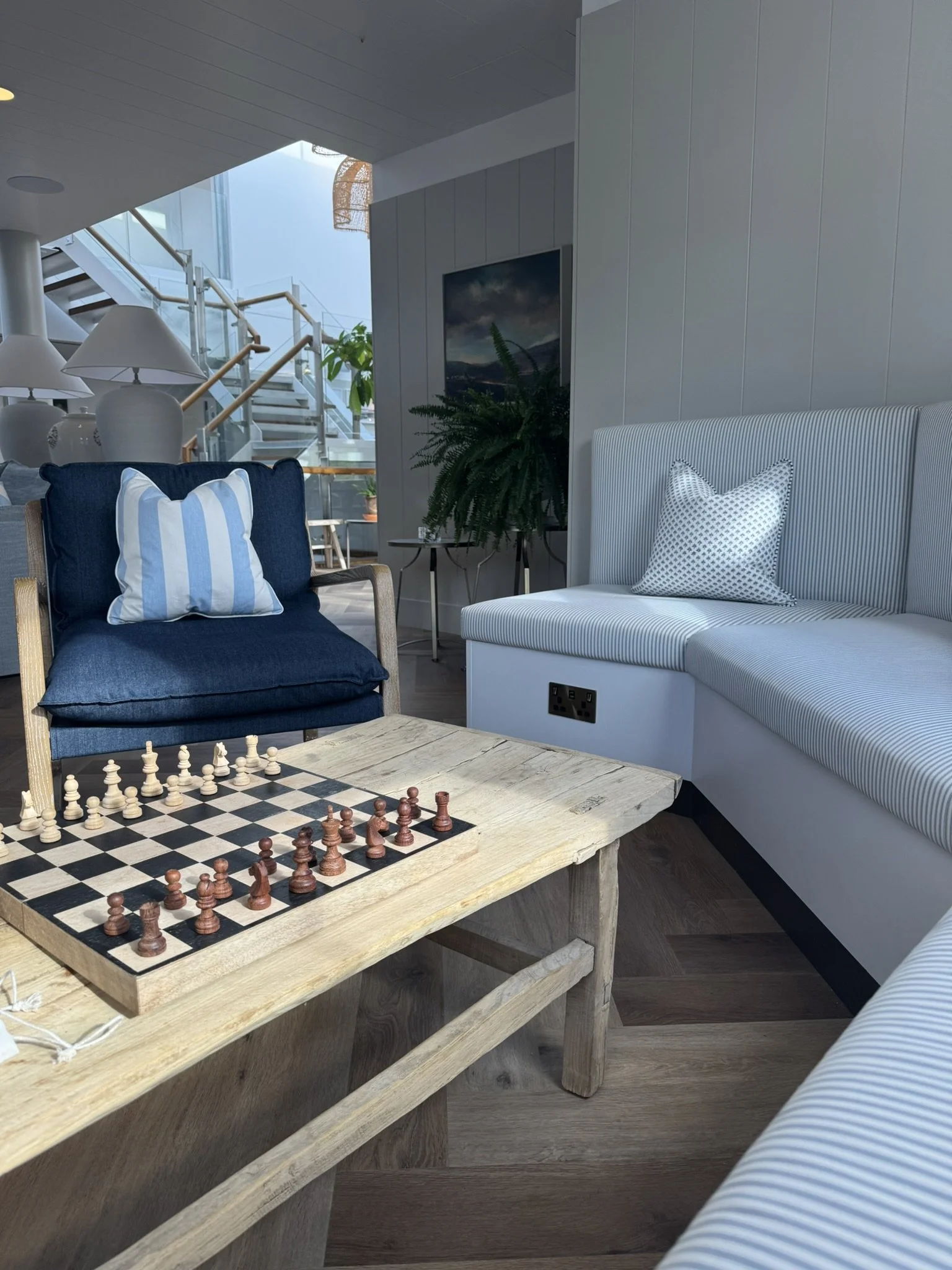 A cozy living room with a chessboard on a wooden table, a blue armchair with a striped pillow, a white striped sofa with another pillow, and a green potted plant near the wall. Stairs and a window are visible in the background.
