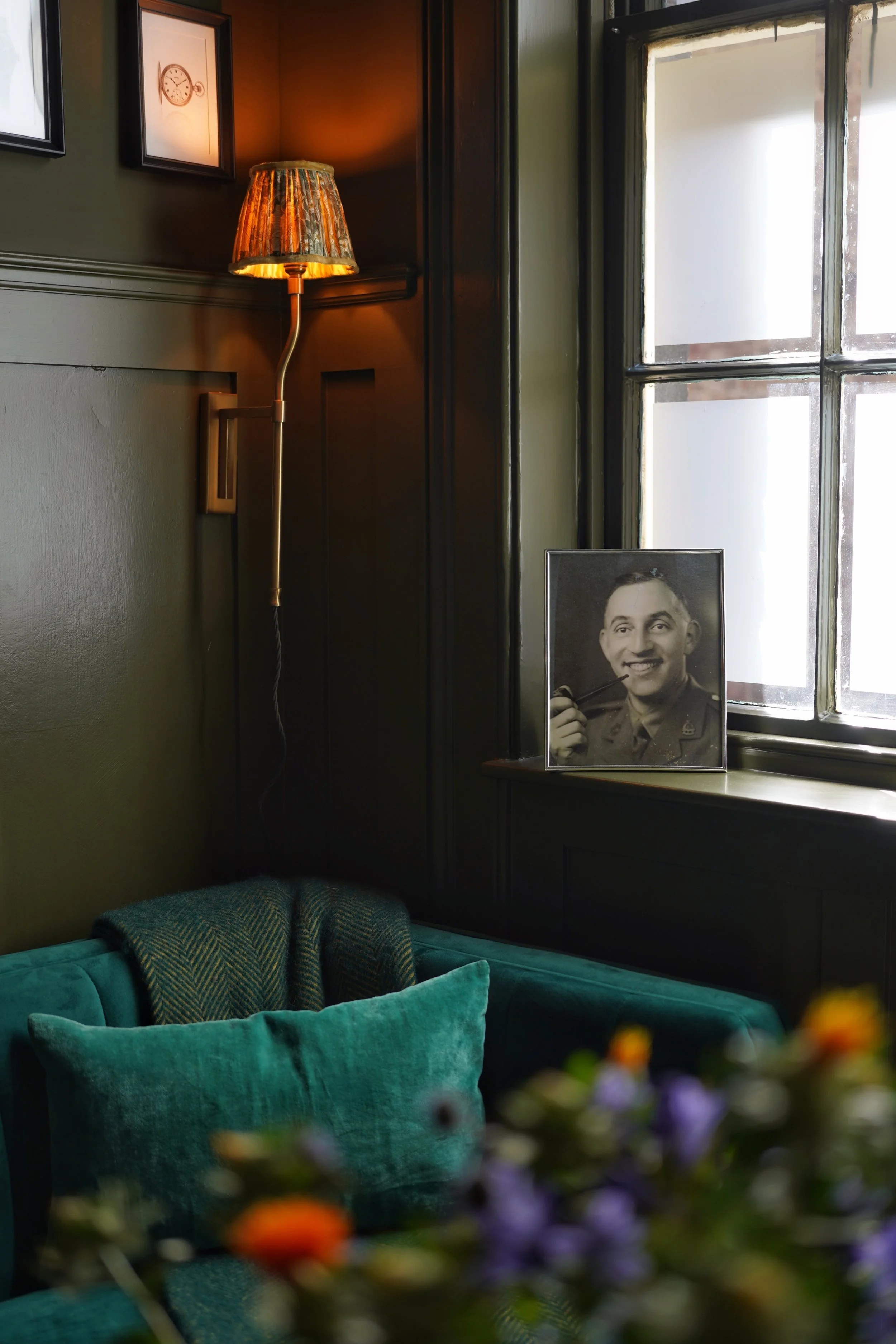 A cozy corner of a room with dark green walls, a vintage-style lamp on the wall, a window letting in natural light, a black-and-white portrait of a man in a military uniform, a teal sofa with cushions, and some flowers in the foreground.