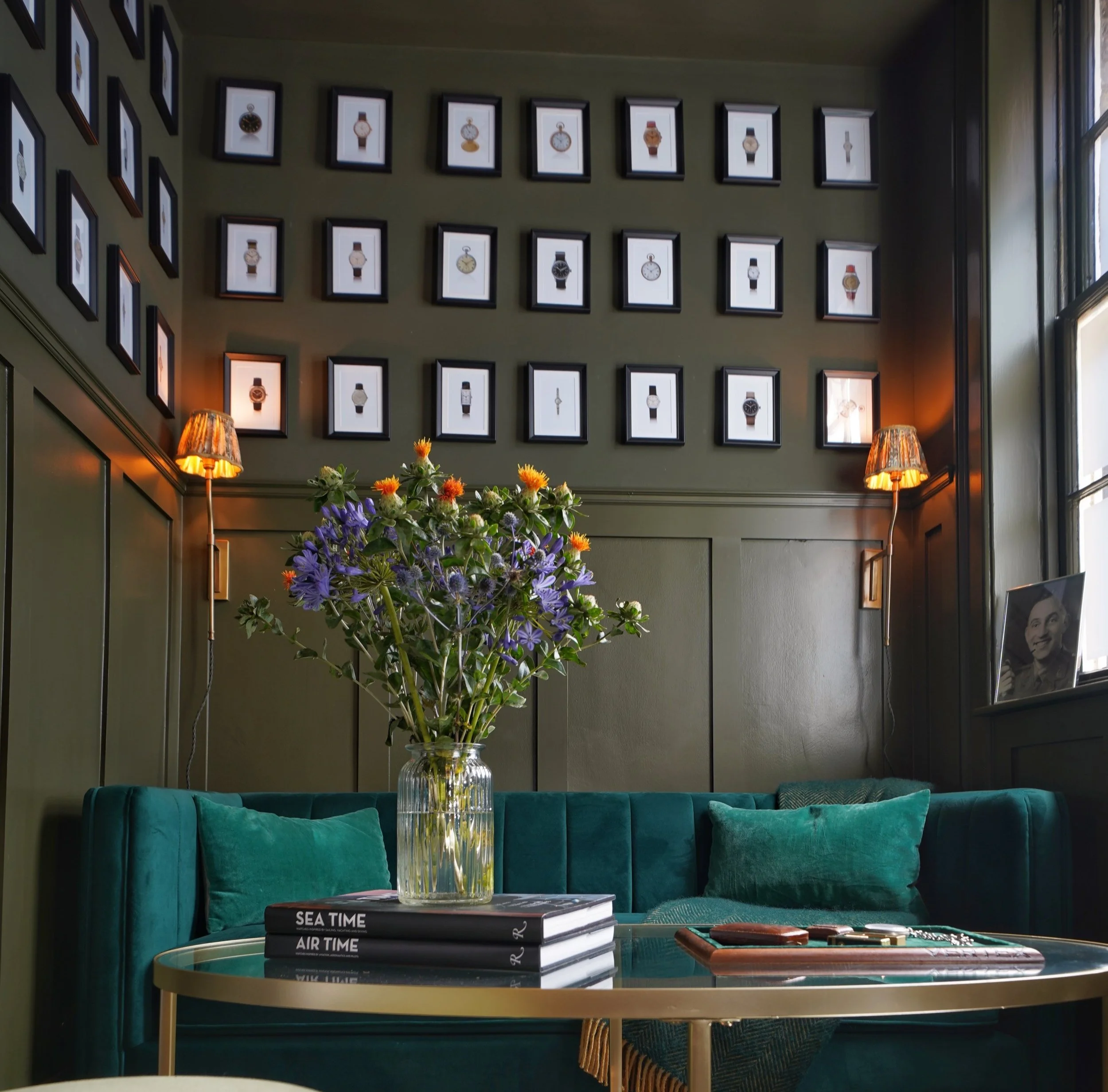 A cozy sitting area with a teal velvet sofa, a round glass coffee table, a vase of purple and orange flowers, and framed watches on the wall. There are two lamps with warm light, and a black-and-white portrait on the window ledge.