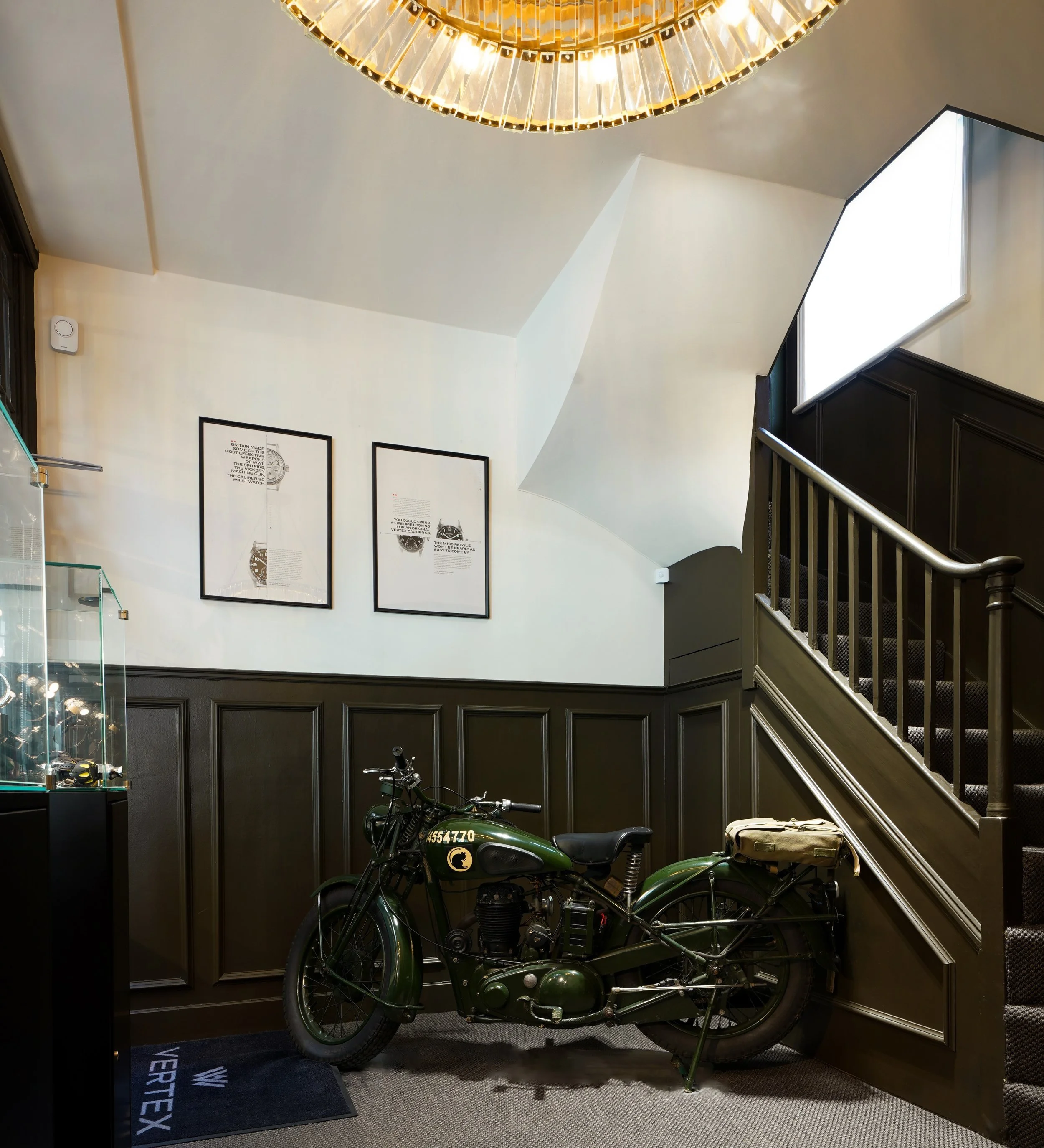 Vintage military motorcycle displayed in a room with dark wood paneling, staircase, and framed artwork on the wall.