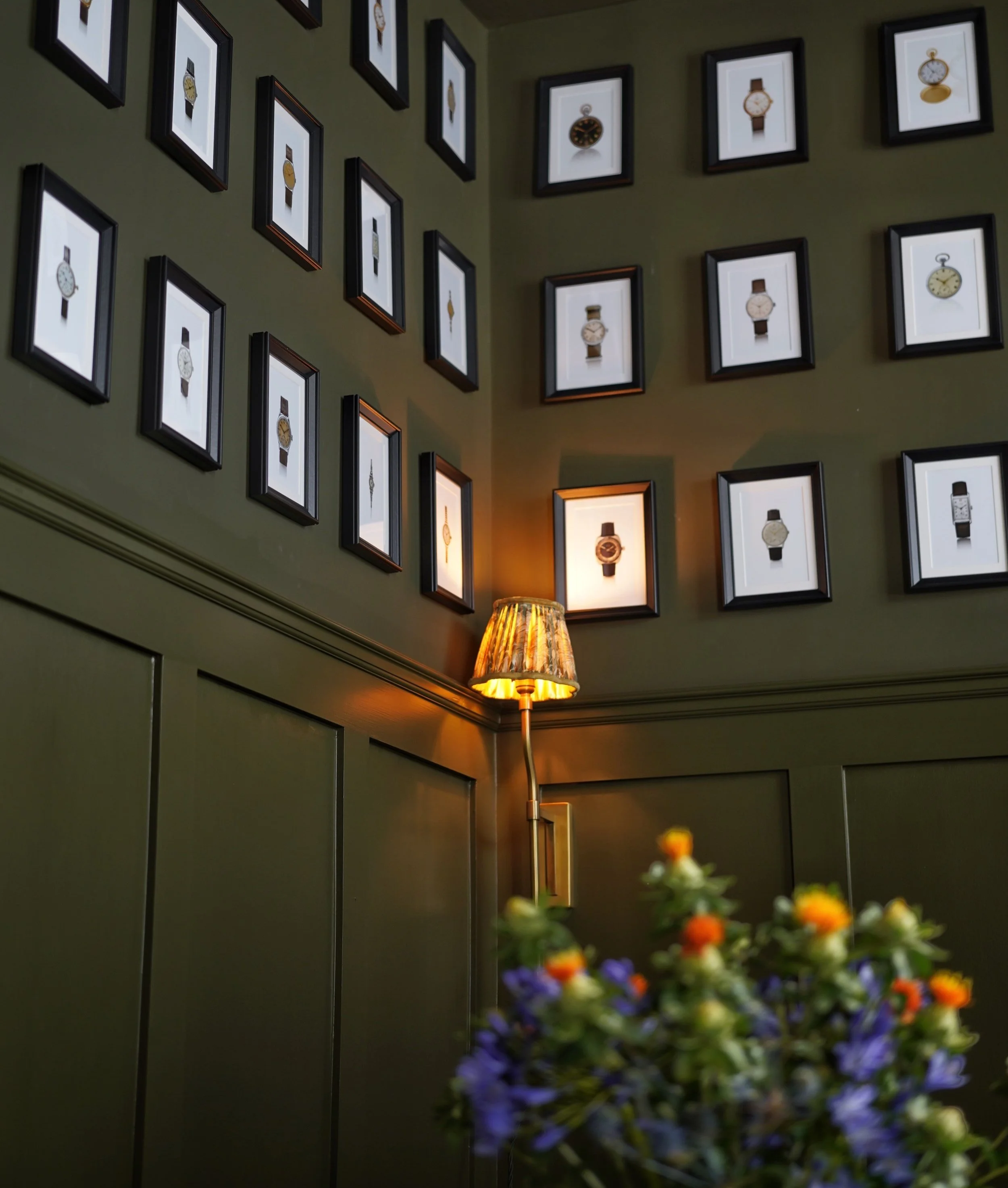 Wall decorated with framed vintage watches, illuminated by a wall-mounted lamp with a floral lampshade, with a vase of colorful flowers in the foreground.