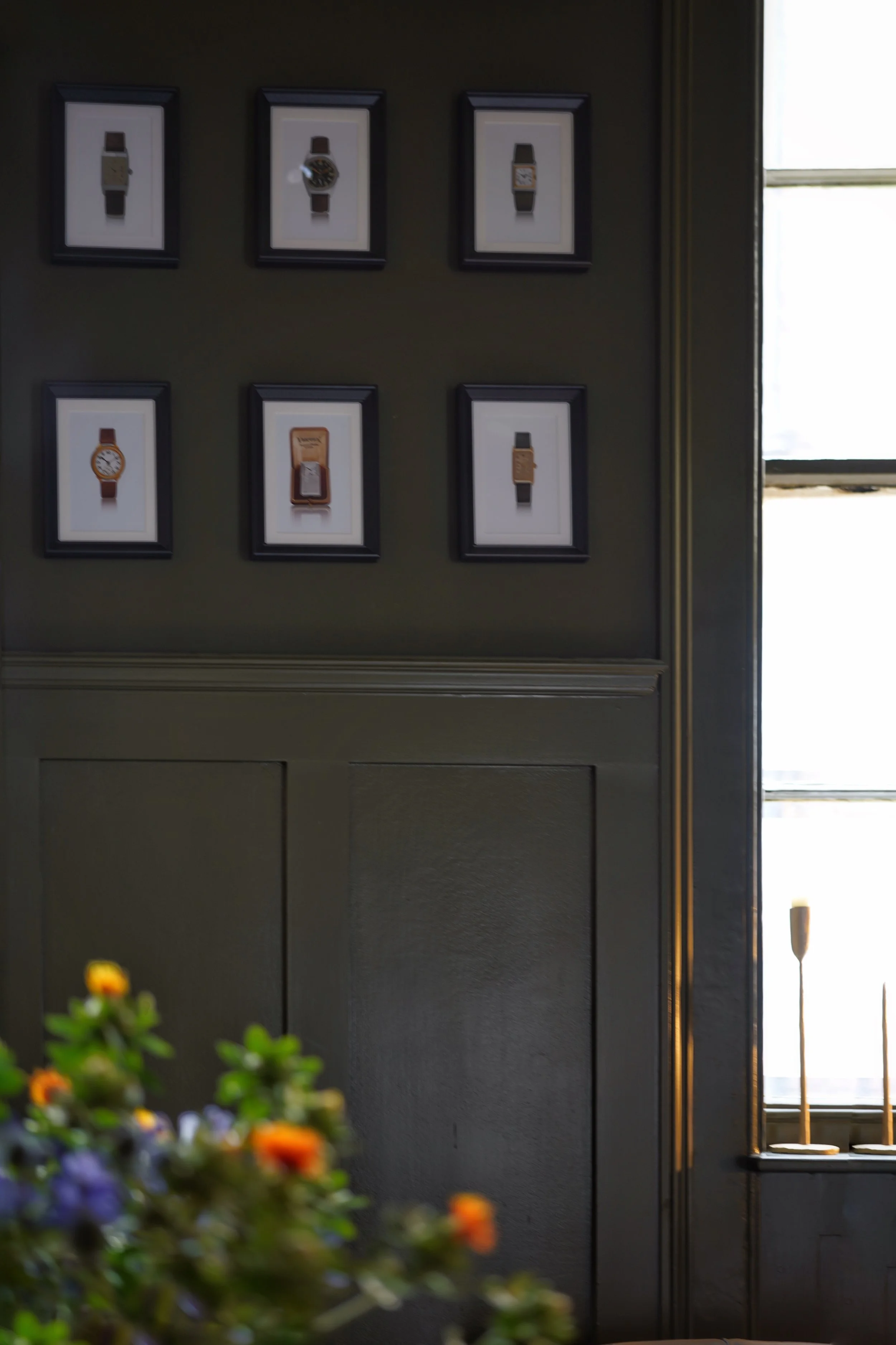 Framed pictures of watches and a small framed image or poster on a dark-colored wall near a window, with a blurred bouquet of flowers in the foreground.