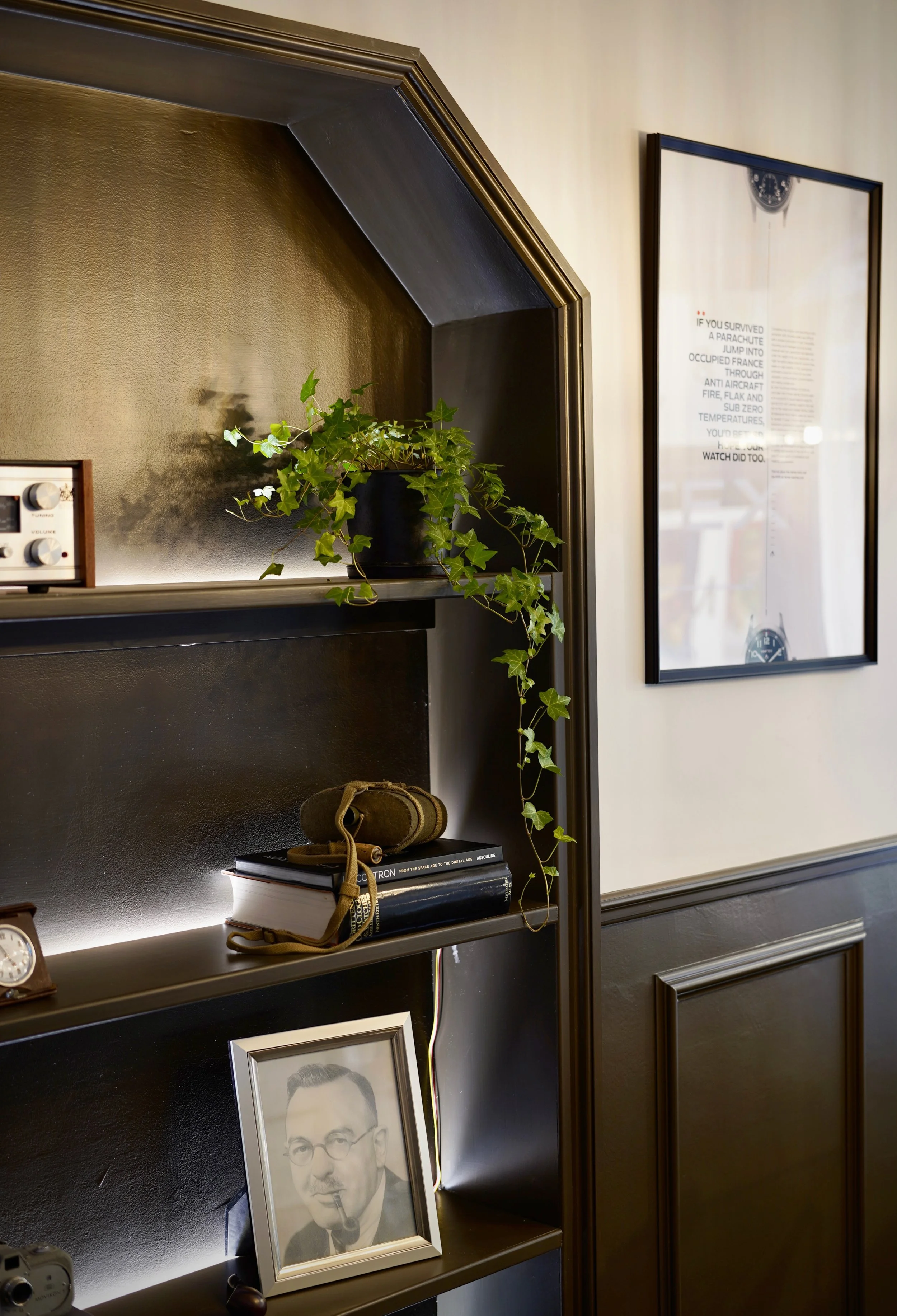 A black bookshelf with a potted ivy plant on top, a book, a wallet, and a framed black-and-white portrait of a man smoking a pipe. Behind the shelf, there is a beige wall with a framed poster or art print and part of a plaque or sign.