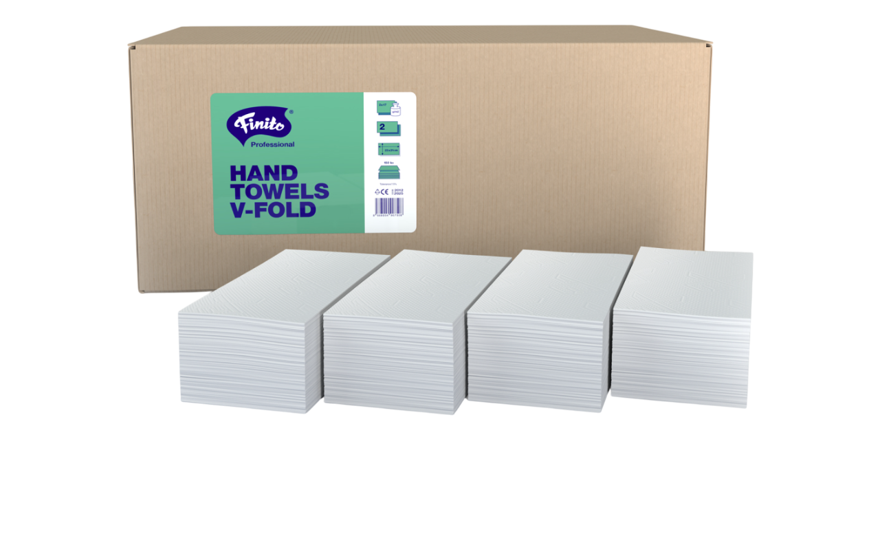Ein Karton mit weißen V-förmigen Handtüchern auf dem Tisch, neben mehreren aufgestapelten Handtüchern, und die Verpackung zeigt das Label "Finito Professional HAND TOWELS V-FOLD".