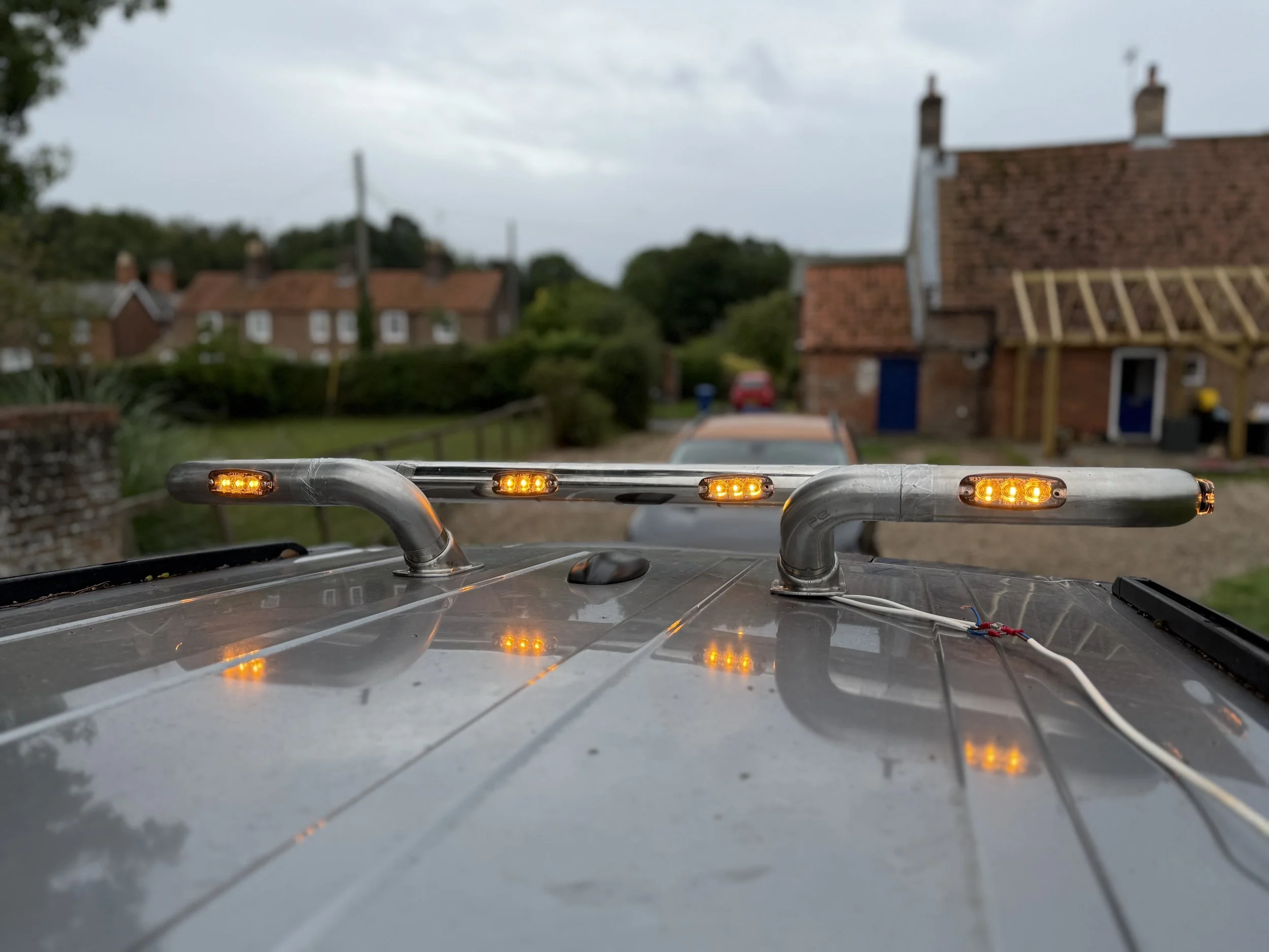 The image shows the roof of a vehicle with four orange indicator lights turned on, with a suburban neighborhood in the background including houses, trees, and a cloudy sky.