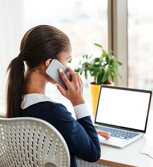 Woman talking on a mobile phone while working on a laptop at a desk in a bright office.