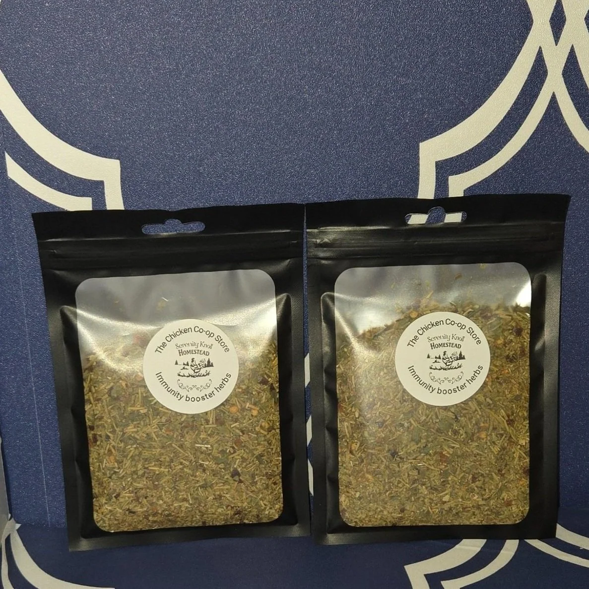 Two resealable black pouches of herbal mixture with white circular labels reading "The Chicken Co-op Store Homestead Community booster herbs" placed on a blue and white patterned surface.