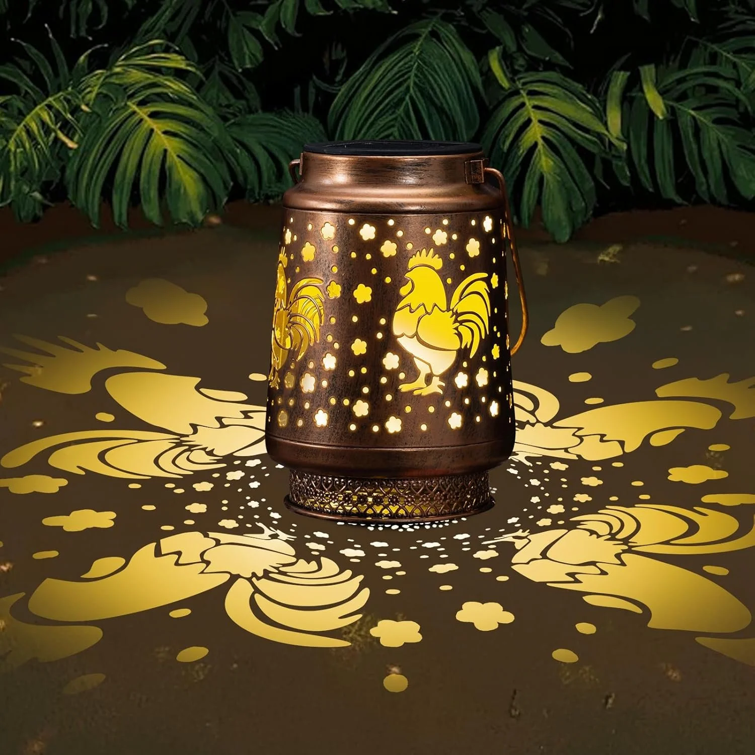 Decorative lantern with rooster cutout illuminating yellow light, casting floral and rooster patterns on surrounding surface, with green leafy plants in background.