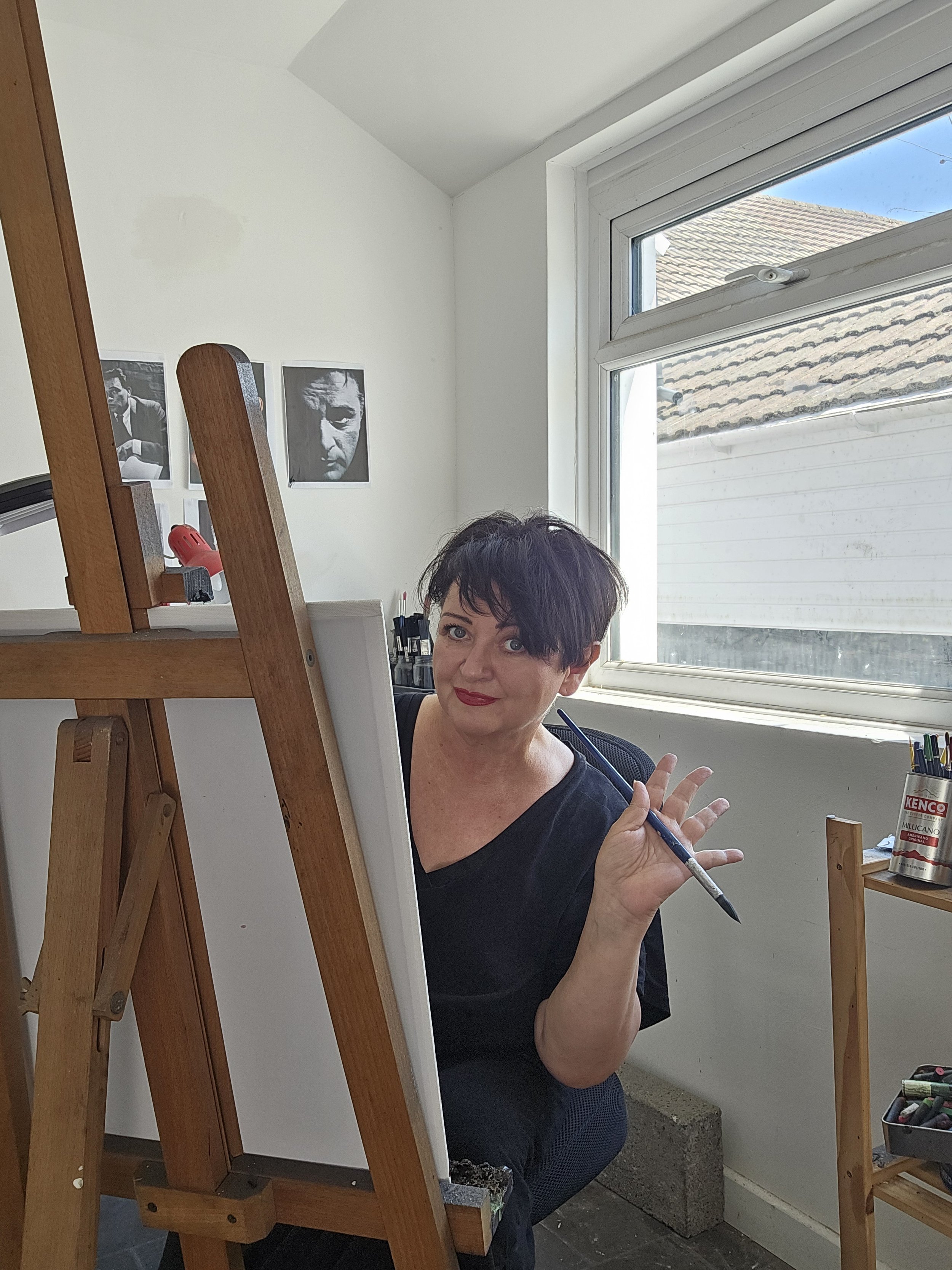 A woman with short dark hair and red lipstick sitting in front of an easel, holding a paintbrush, in a bright room with a window and black-and-white photographs on the wall.