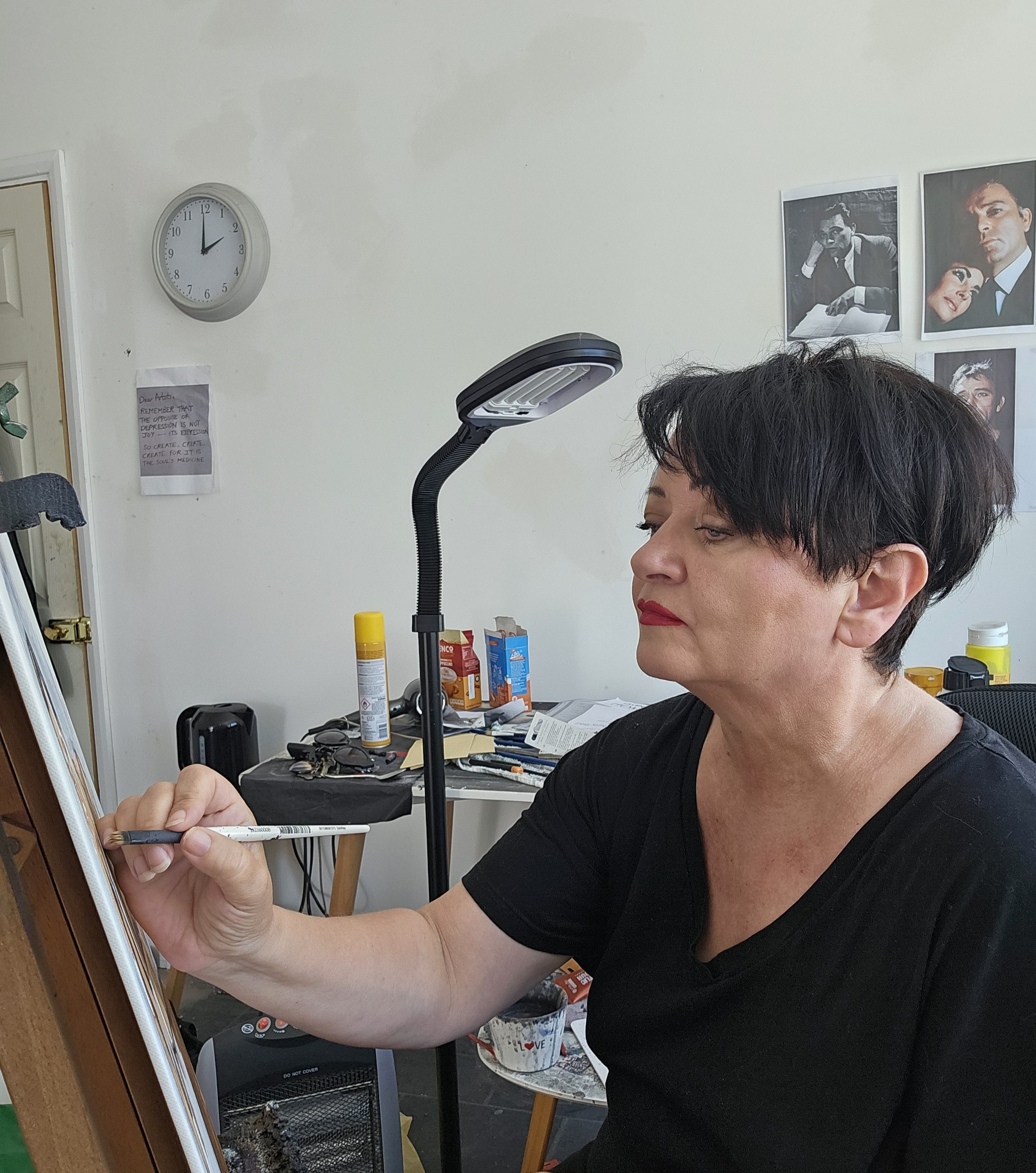 A woman with short dark hair and red lipstick is drawing on a canvas or paper, seated at a desk with art supplies and clutter. The room has a wall clock showing 12:00, a standing lamp, some photographs, and various items on the desk.