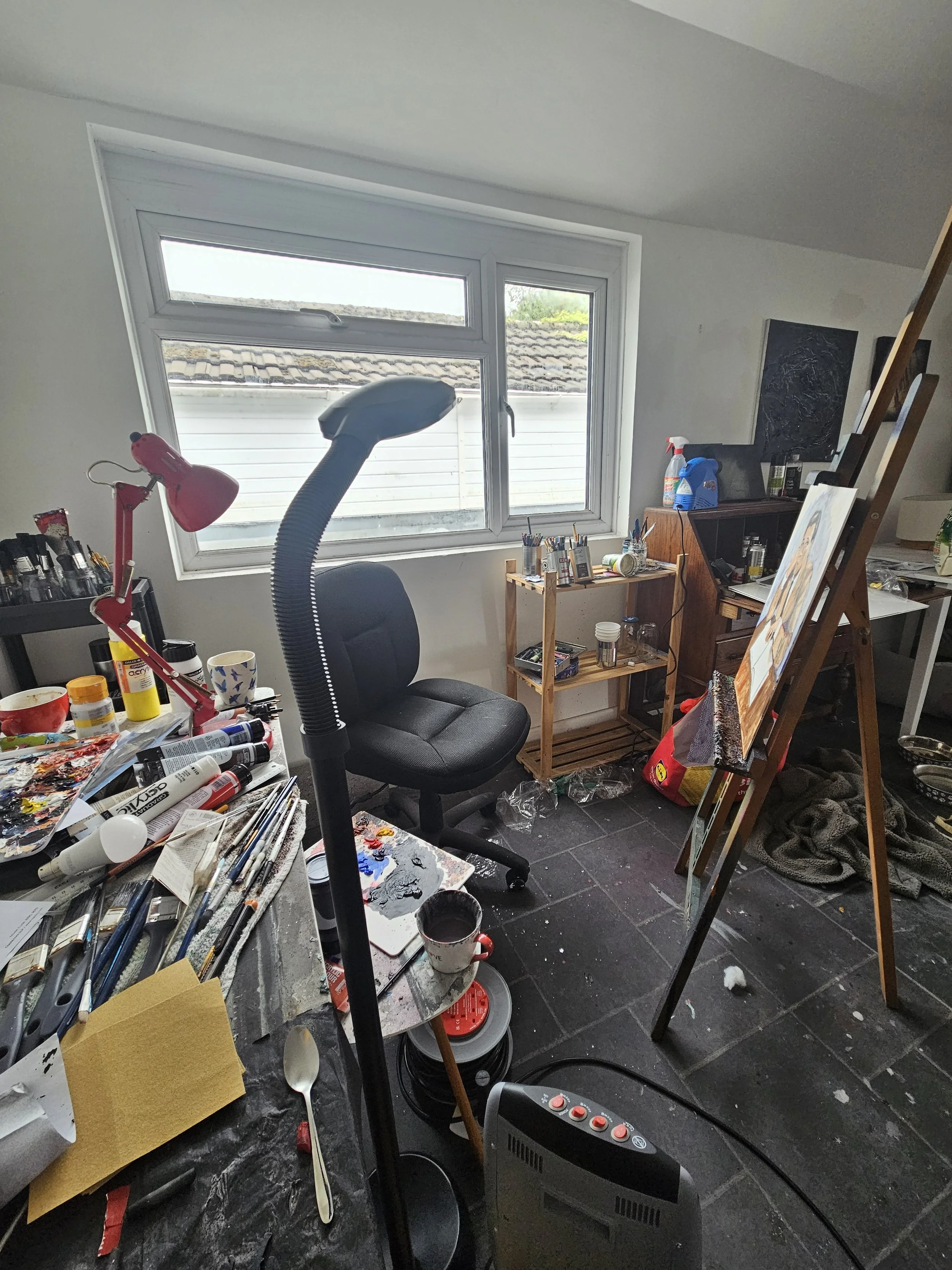 Artist's workspace with painting supplies, a canvas on an easel, a black office chair, and scattered art tools on a cluttered table, with a window in the background.