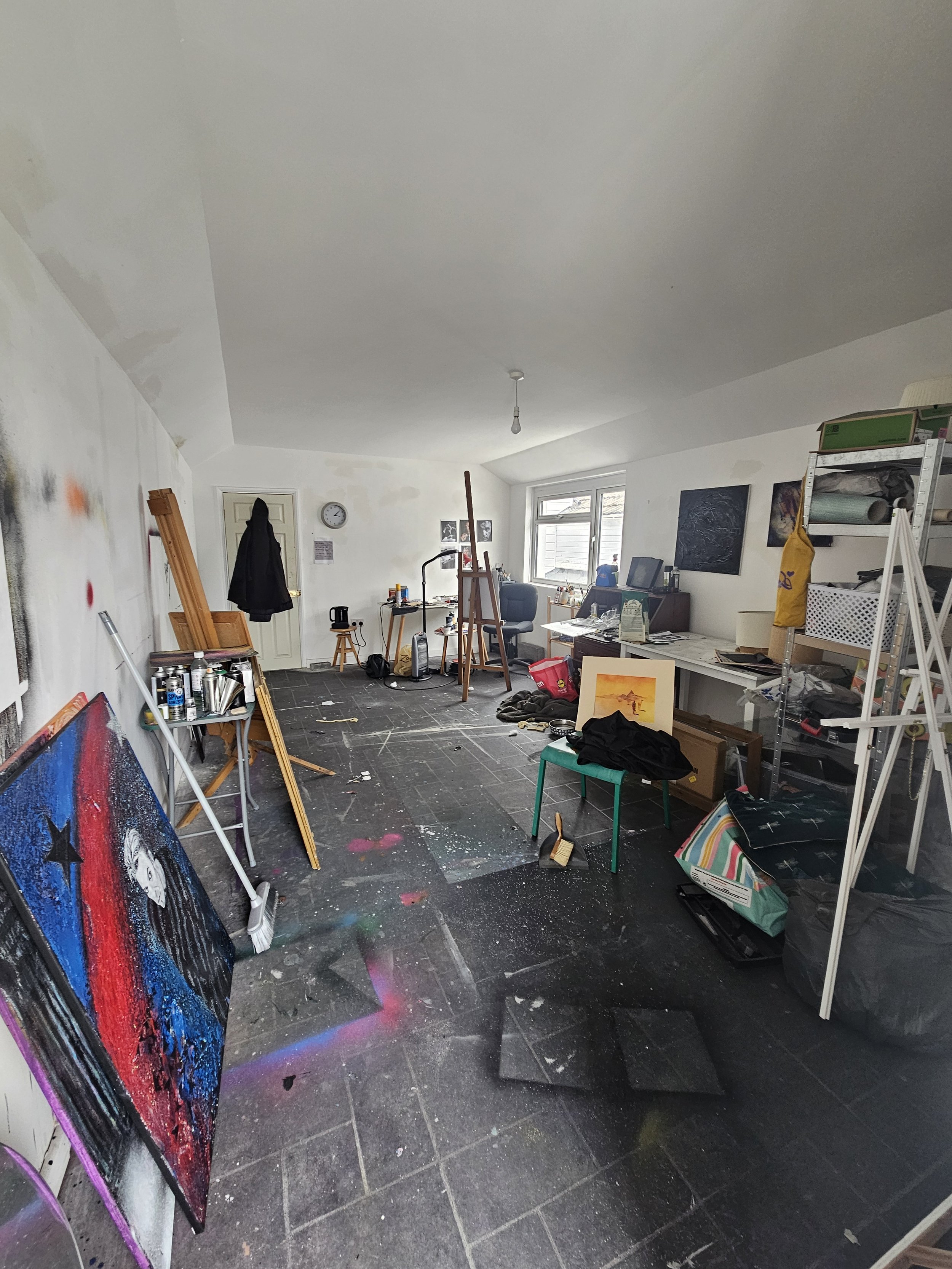 A cluttered art studio with paintings, tools, and supplies scattered around, near a window and a door.
