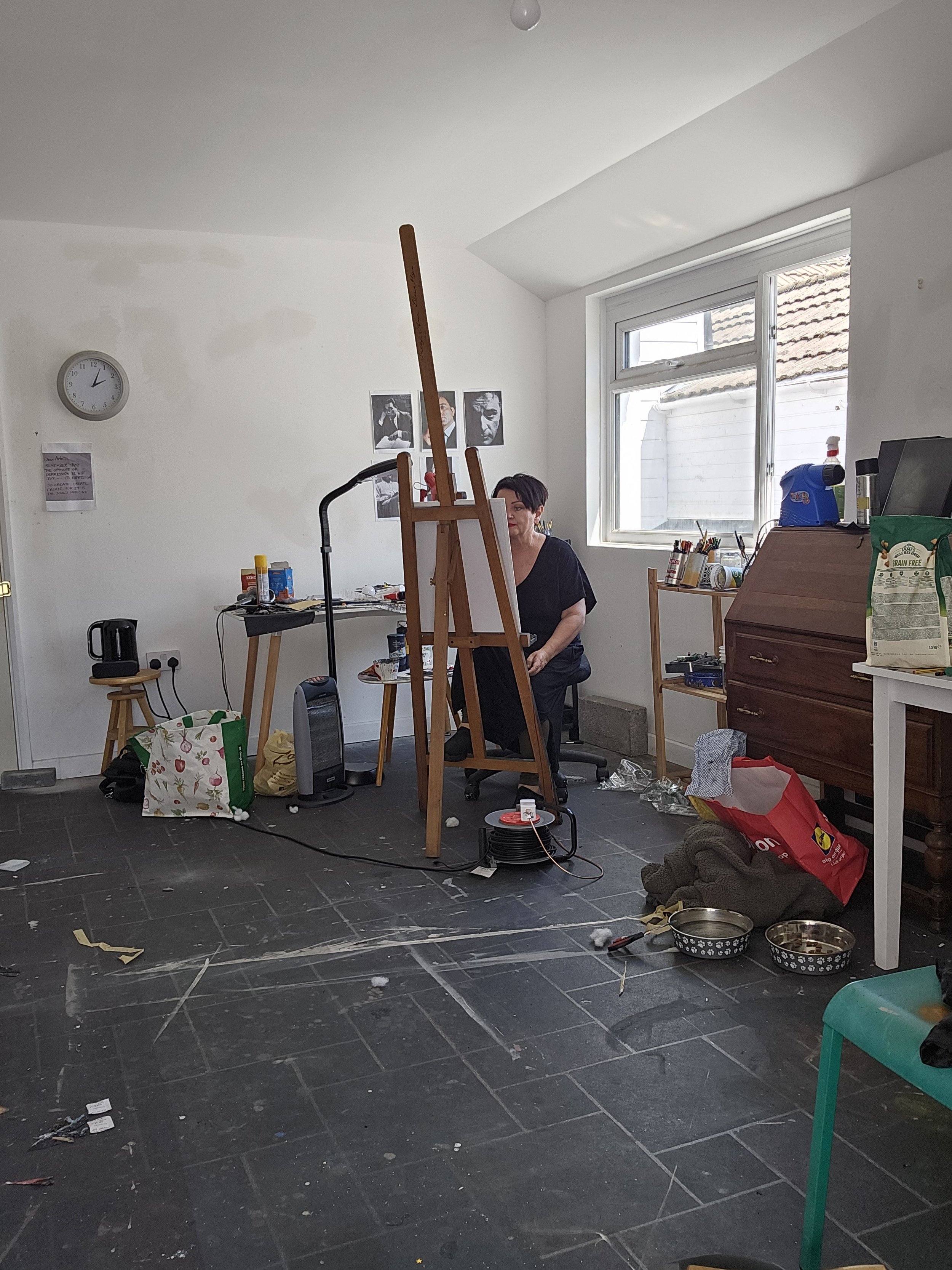 Room with a woman sitting at an easel, surrounded by art supplies, with a window to the outside, a clock on the wall, and various scattered items on the floor.