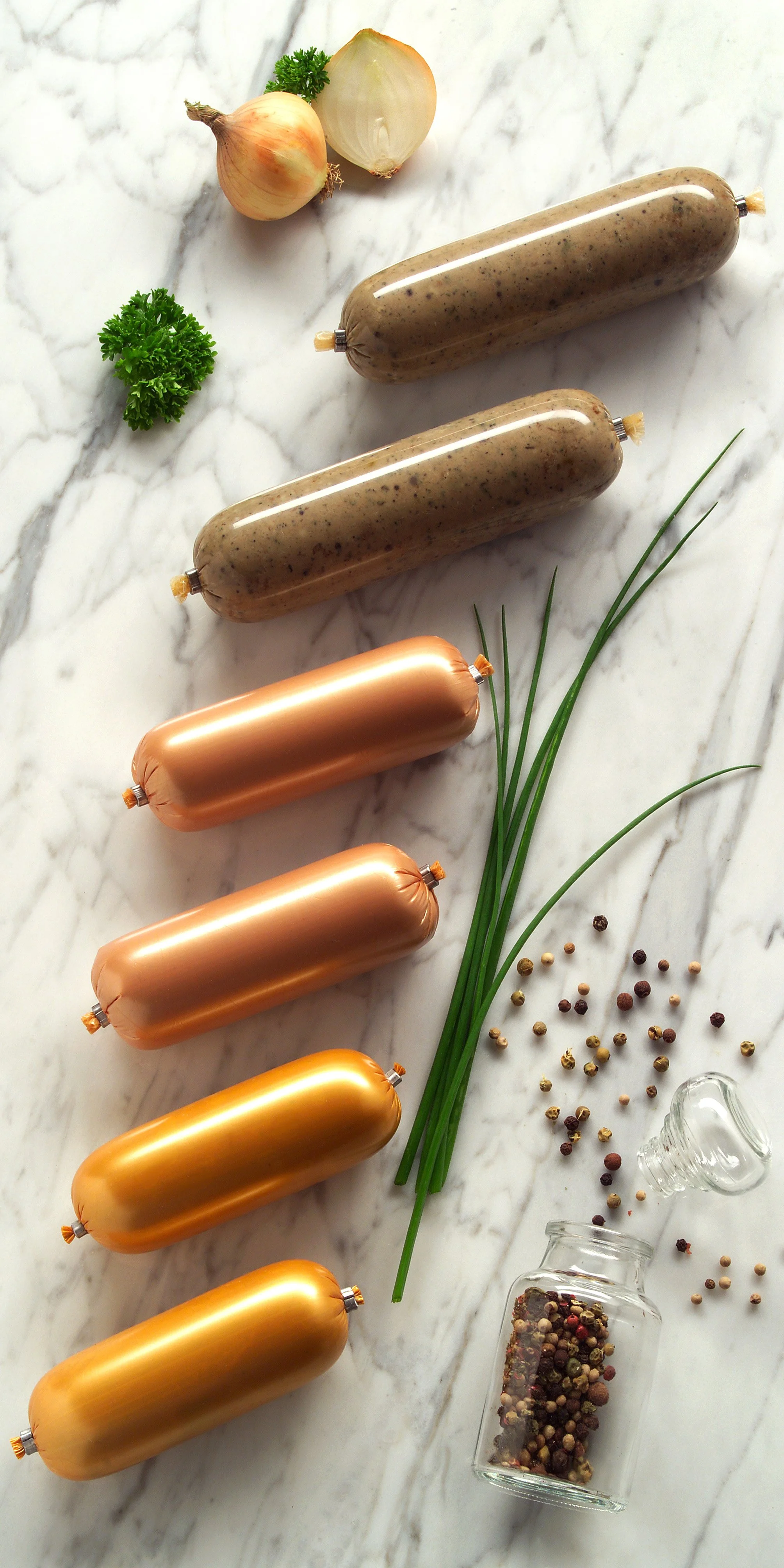 Siete salchichas en diferentes colores y tamaños, cebolla, perejil, cebolla de verdeo, sal de pimienta en salsera de vidrio y variedad de pimientas en frascos de vidrio sobre una superficie de mármol