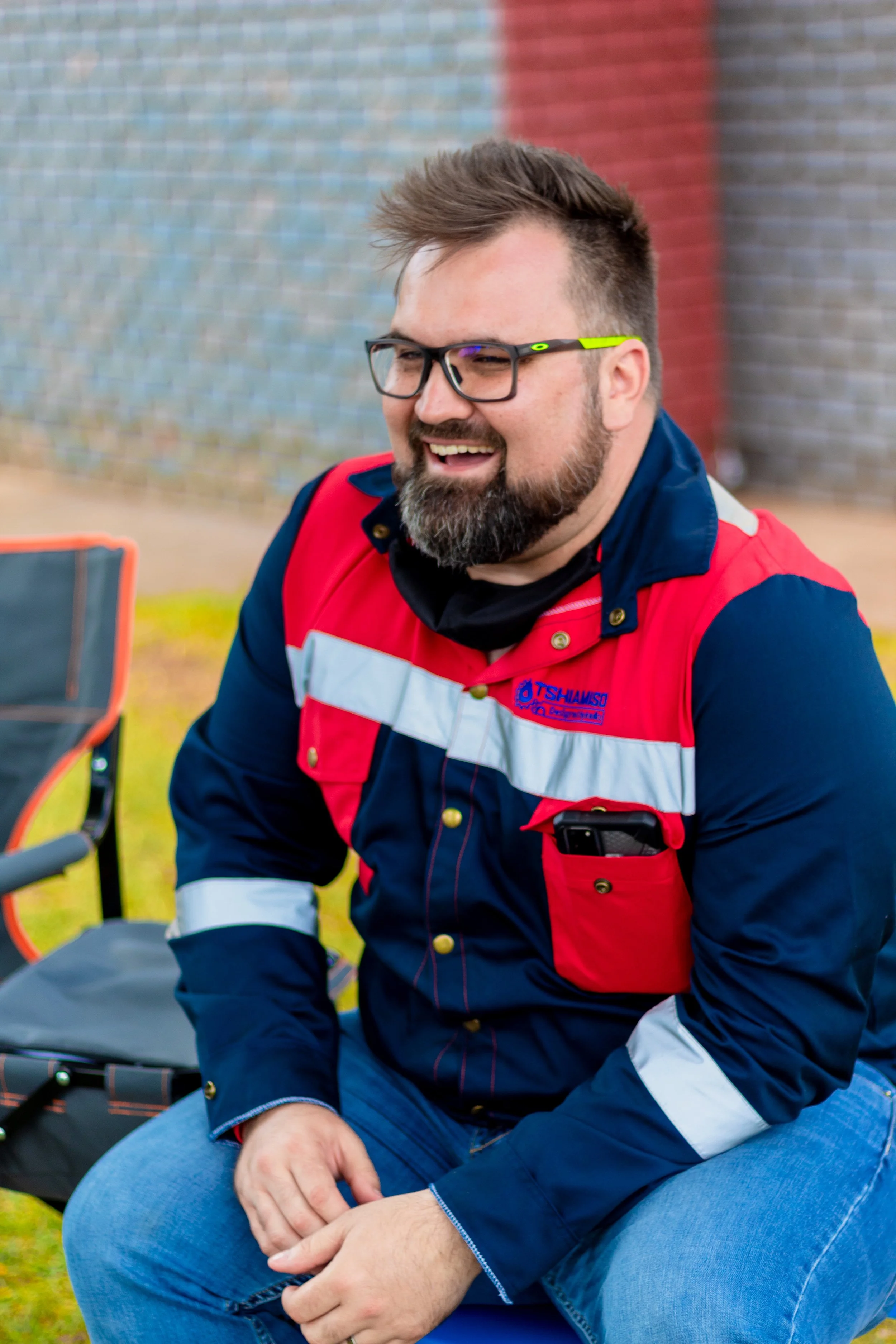 A man with glasses, a beard, and a mustache, smiling and sitting outdoors. He's wearing a red, navy, and white uniform shirt with a logo "Tshamiso" and has a phone in his shirt pocket, with a folding chair beside him.