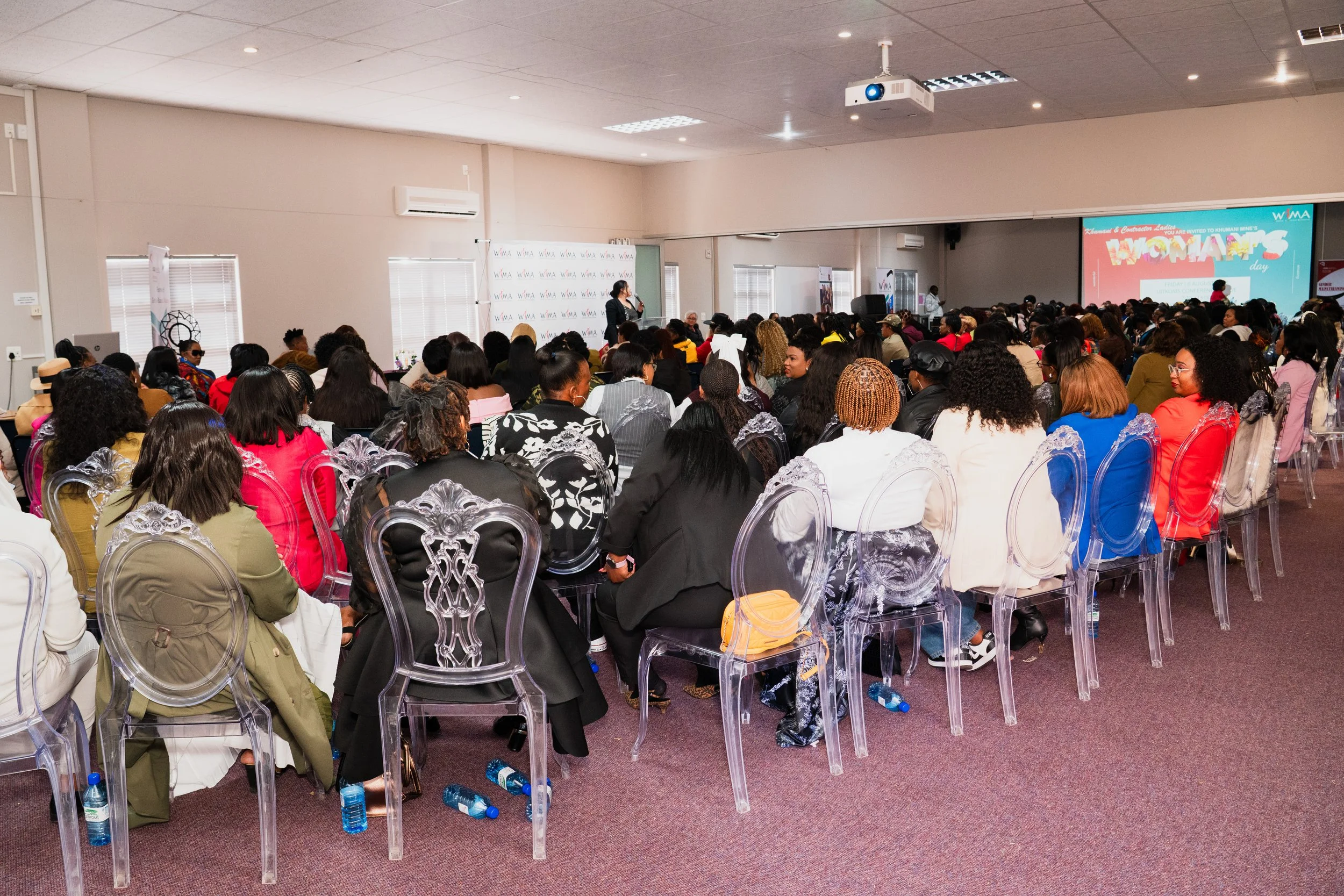 A large conference room filled with a diverse crowd of women attending an event. They are seated in transparent chairs, facing a stage with a woman speaker and a large screen displaying a presentation.