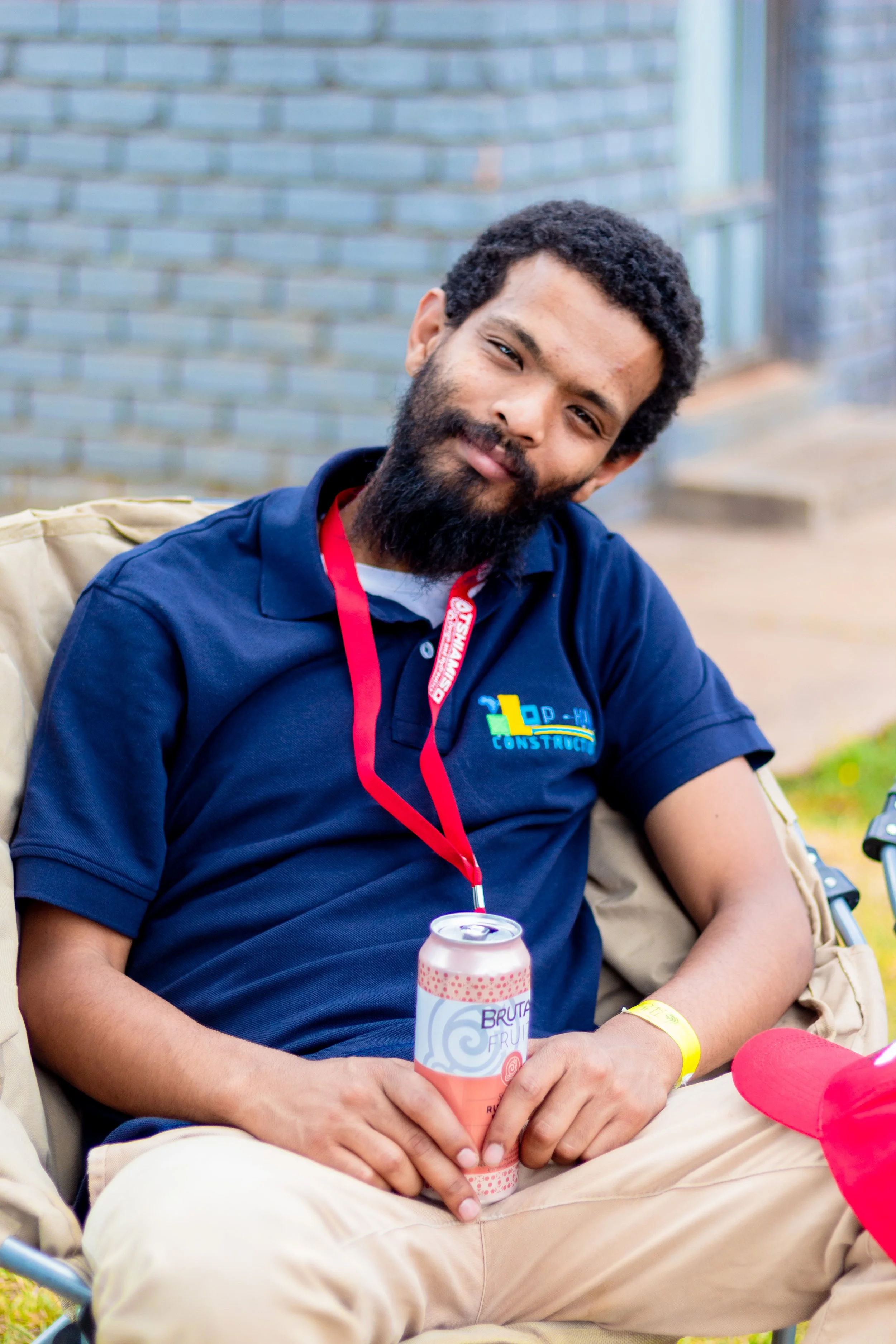 A smiling man with a beard and short curly hair, wearing a dark blue polo shirt with a construction logo, sitting outdoors on a beige folding chair, holding a canned beverage, with a red event badge around his neck.