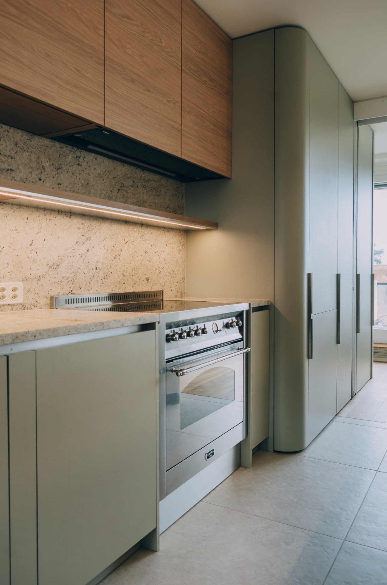 Cuisine moderne avec meubles verts, comptoir en pierre, four en acier inoxydable, armoires en bois foncé, et grande fenêtre laissant entrer la lumière naturelle.