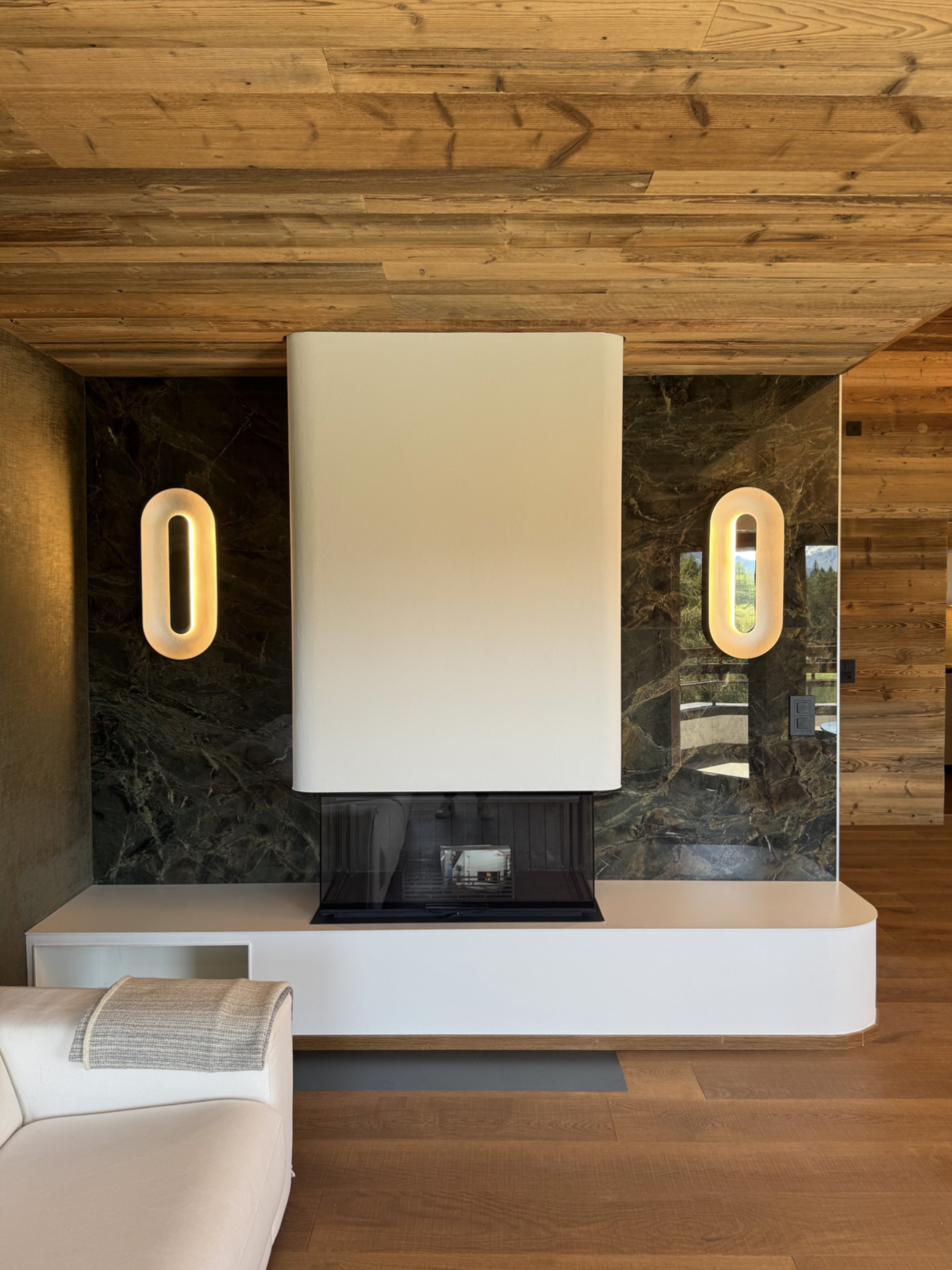 Cheminée moderne encastrée dans un mur en marbre avec deux lampes ovales murales éclairantes de chaque côté, dans une pièce avec plafond en bois et mobilier beige.
