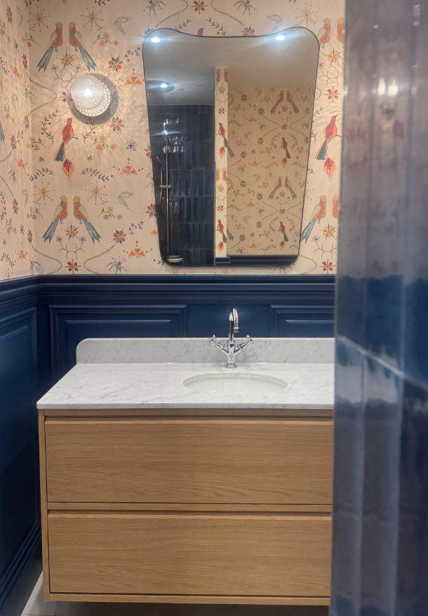 Vue partielle d'une salle de bain avec un miroir rectangulaire, un lavabo avec comptoir en marbre, un meuble en bois clair, et un mur décoré avec du papier peint à motif de oiseaux et de fleurs, ainsi qu'une applique lumineuse au plafond.
