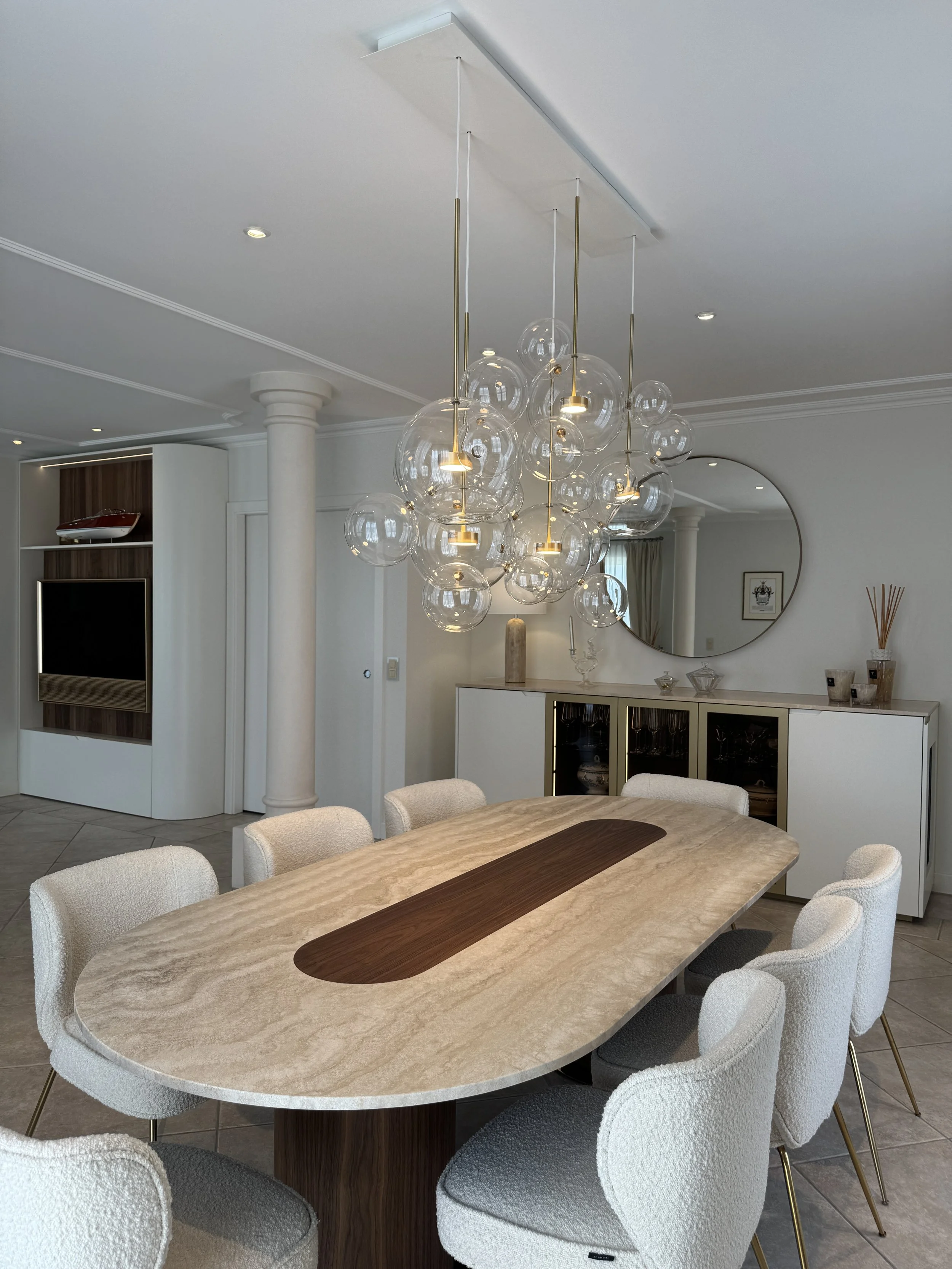 Salle à manger avec une table en bois beige et un centre en bois foncé, entourée de chaises creme avec des jambes en métal doré, suspendue à un plafond une suspension en verre avec plusieurs bulles et ampoules, dans une pièce lumineuse avec miroir ro