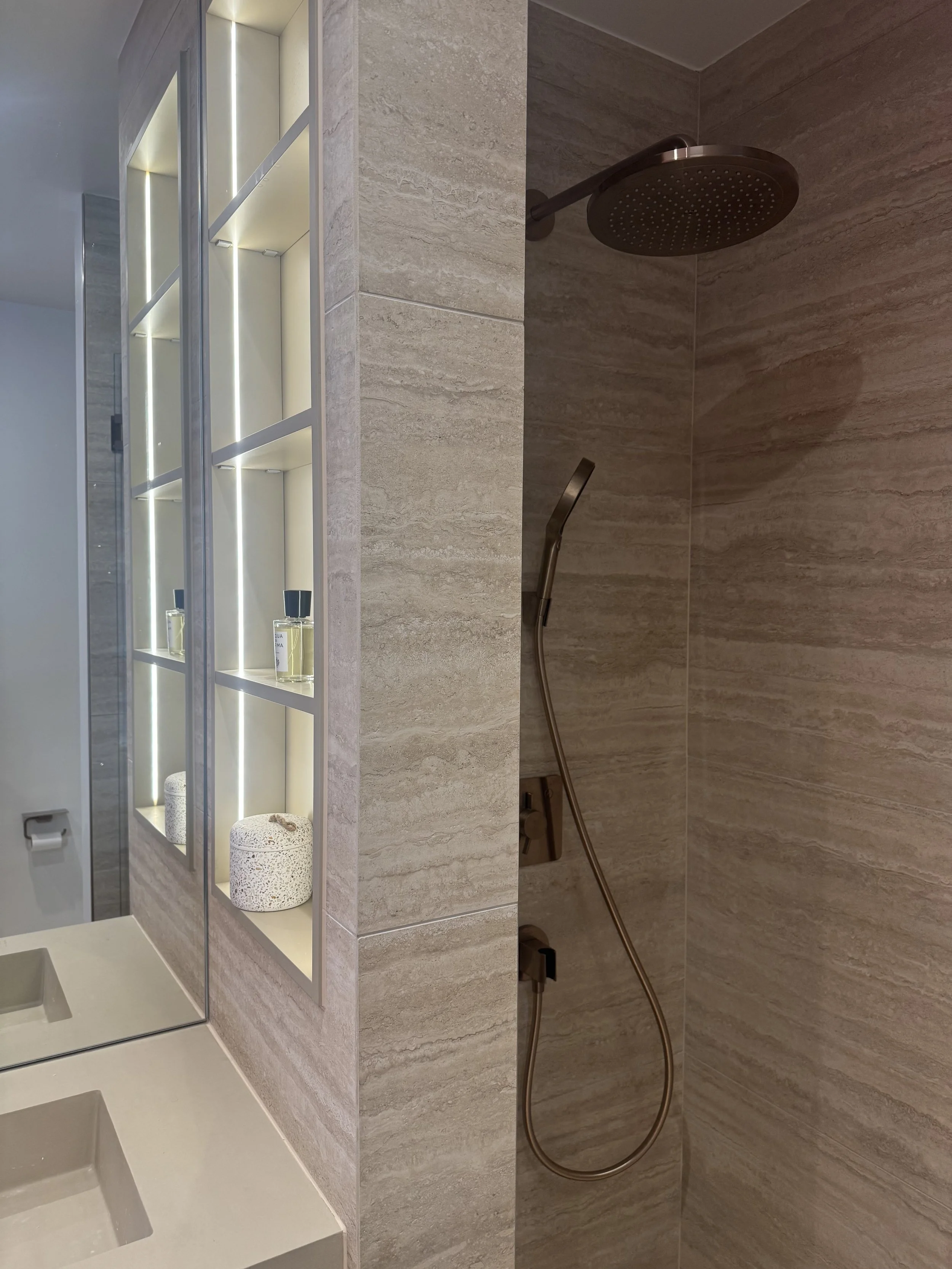 Salle de bain moderne avec douche à pluie, étagères intégrées avec produits de toilette, et lavabo avec miroir.