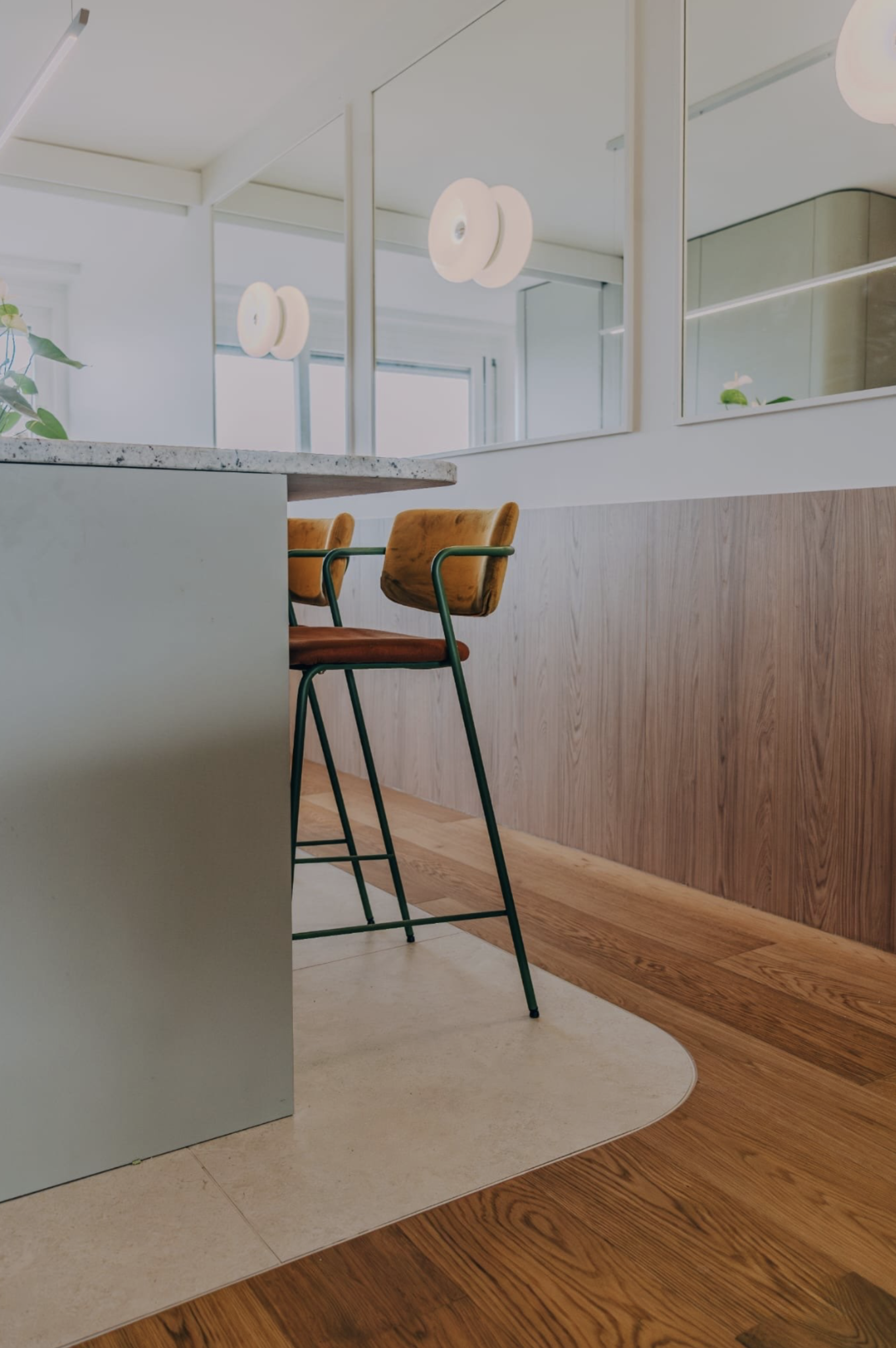 Coin de cuisine avec un comptoir en granit, deux chaises modernes en velours orange et vert, grande fenêtre, miroirs, éclairage suspendu, sol en bois et tapis beige.