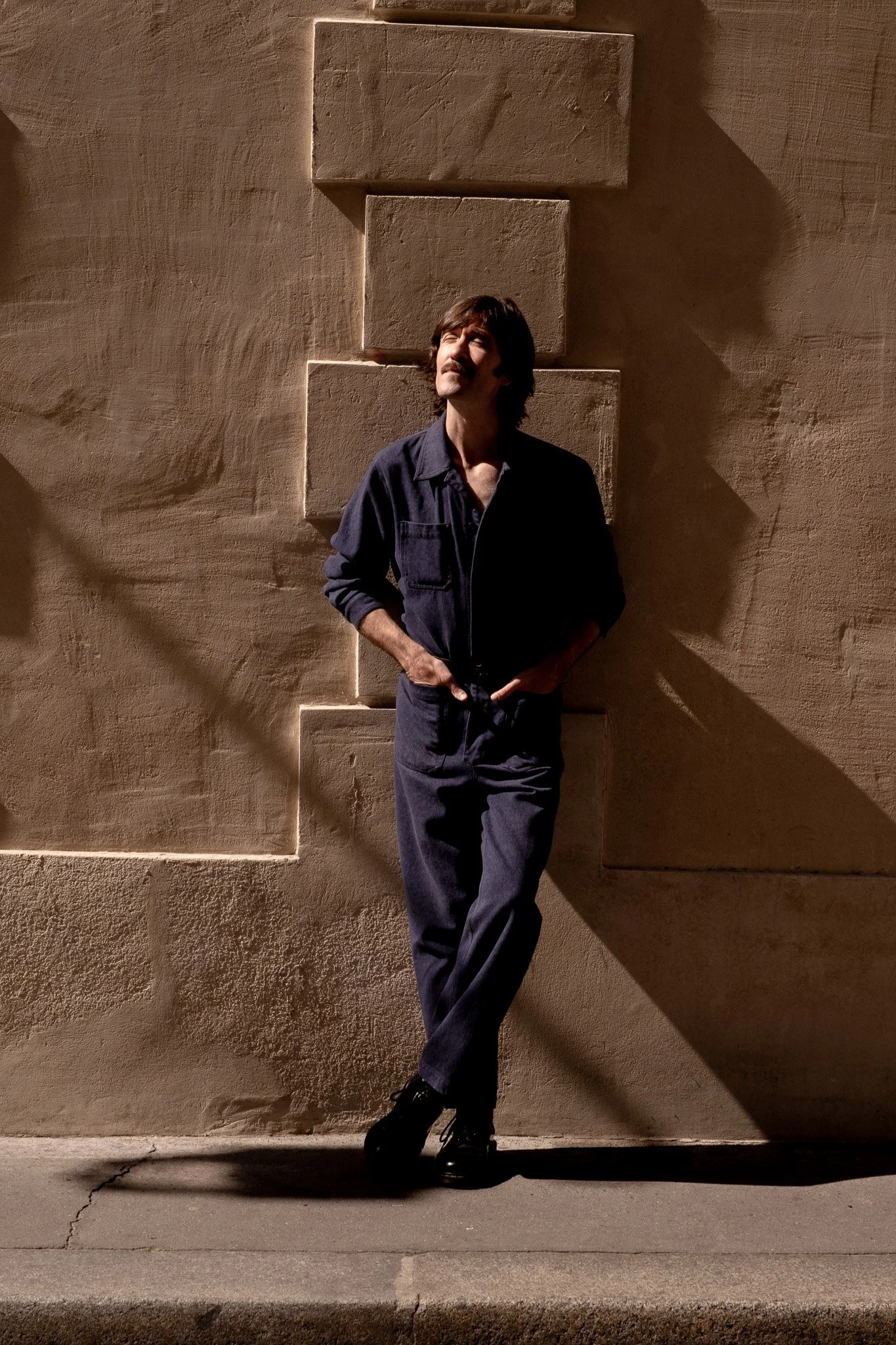 Un homme debout contre un mur en pierre, avec des ombres longues, portant une tenue sombre, pose détendue.