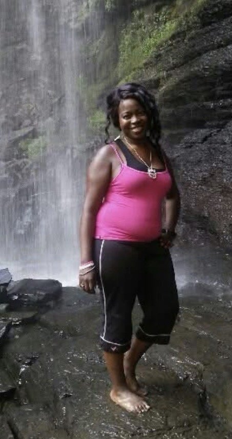 A woman standing barefoot on rocks near a waterfall in a natural outdoor setting, smiling at the camera.