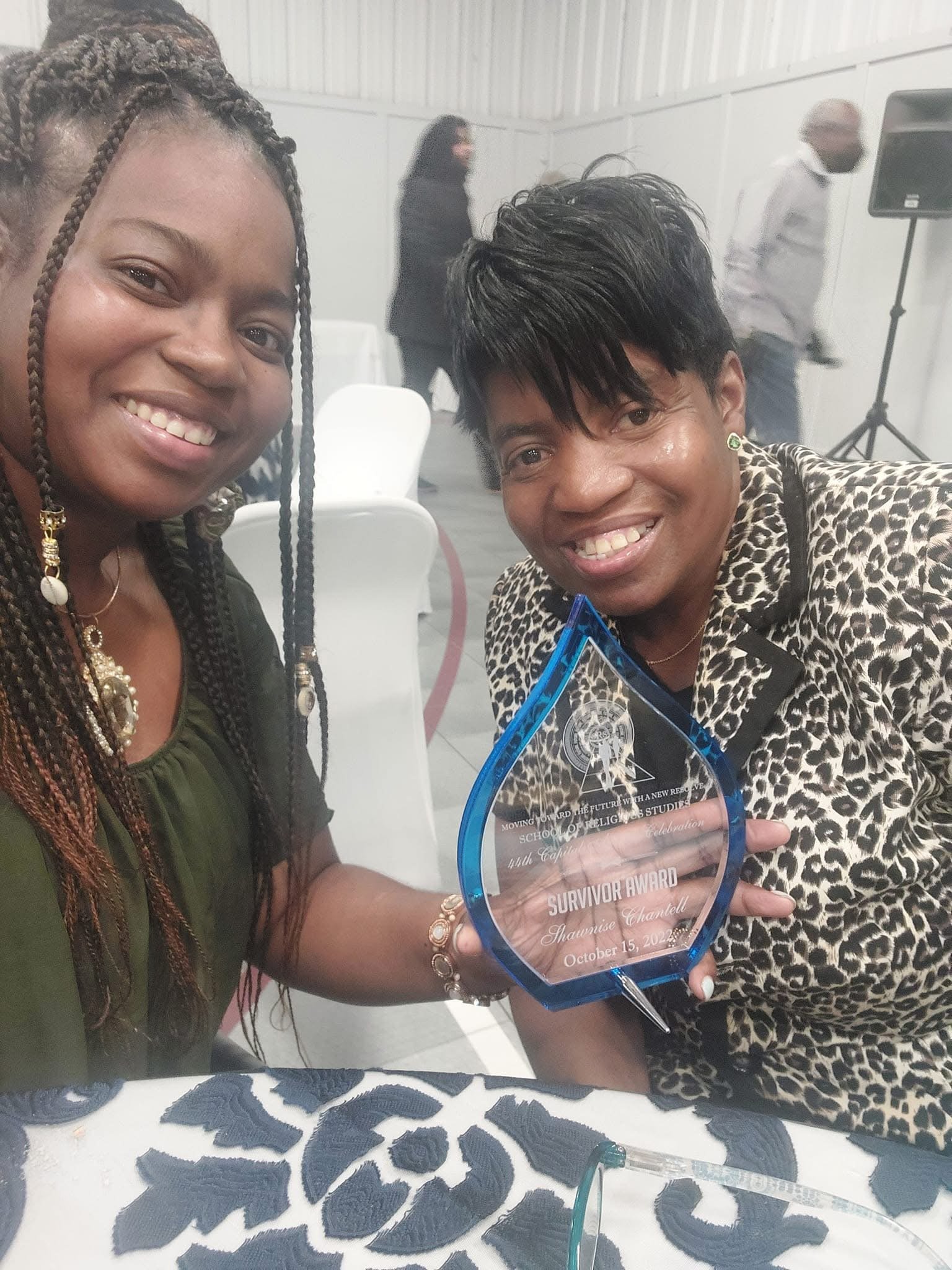 Two women smiling at an award ceremony, one holding a 'Survivor Award' plaque, seated at a table with a patterned tablecloth, with other attendees and a speaker in the background.