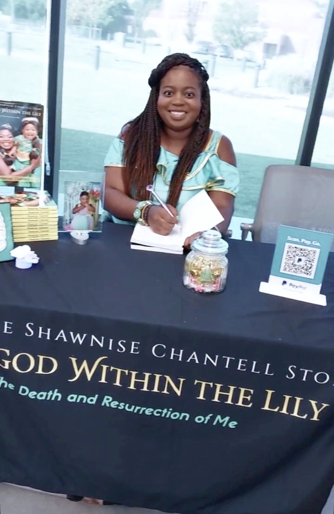 A woman with braided hair sitting at a table signing a book during a book signing event, with a display of her book titled 'God Within the Lily: The Death and Resurrection of Me' by Shawniise Chantell Storm.