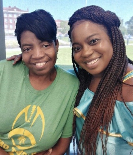 Two women smiling and posing for a selfie outdoors, one wearing a green shirt and the other in a light blue dress with gold accents.