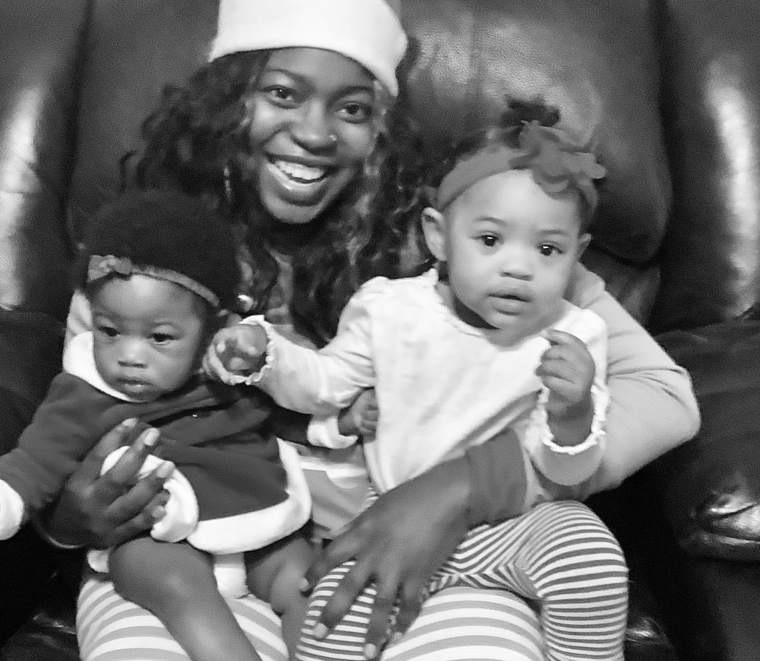 A woman wearing a Santa hat sits on a leather chair, smiling, with two young children on her lap. One child is wearing a dark outfit with a headband, and the other is dressed in a light top and striped pants, looking at the camera.