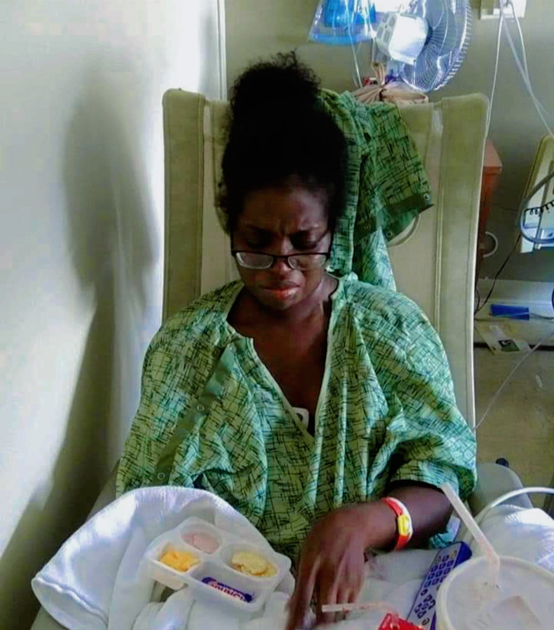 A woman in a hospital bed, wearing a green hospital gown, checking her blood sugar with a glucometer. She has glasses and a healthcare bracelet on her wrist. Medical equipment is visible in the background.