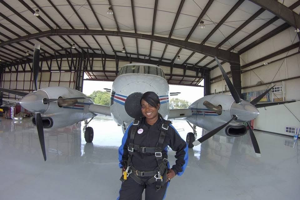 A woman in aviation gear standing in front of a small propeller airplane inside a hangar.