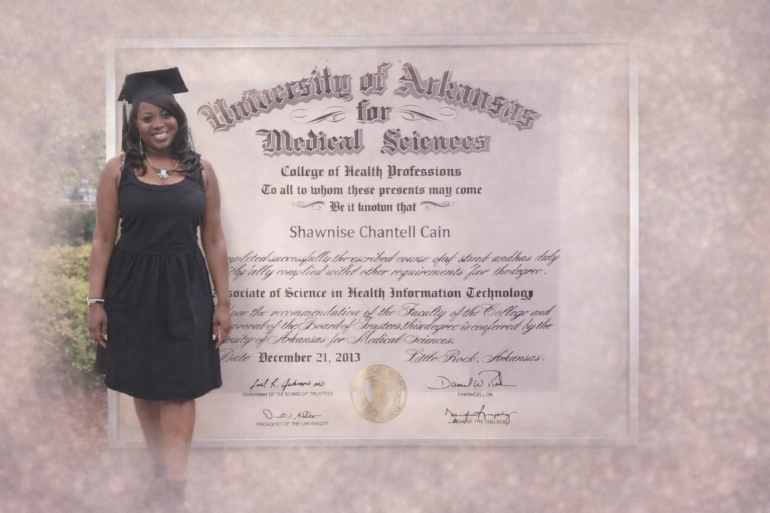 A young woman in a black dress and graduation cap standing next to a diploma from the University of Arkansas for Medical Sciences, College of Health Professions, awarded to Shawneise Chantell Cain.